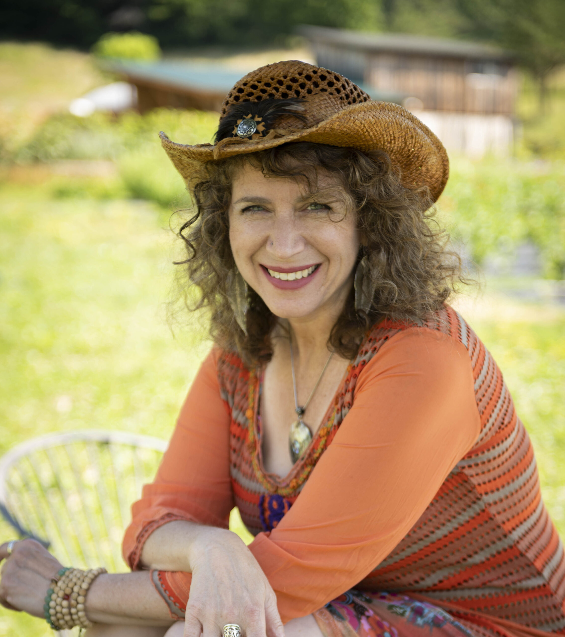 Junie Moon with curly hair wearing a woven hat and an orange top smiles outdoors in a garden setting, embodying the power of self-love.