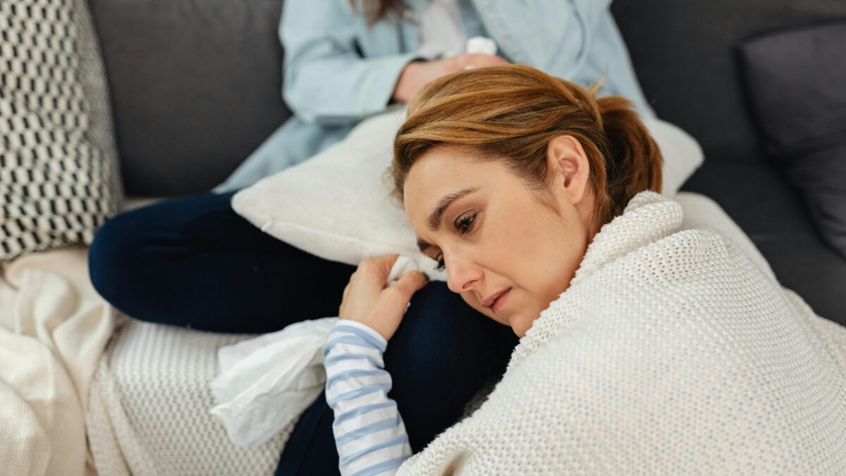 A woman wrapped in a blanket looks contemplative as she rests her head on a sofa cushion, capturing the essence of midlife support, with another person partially visible in the background.