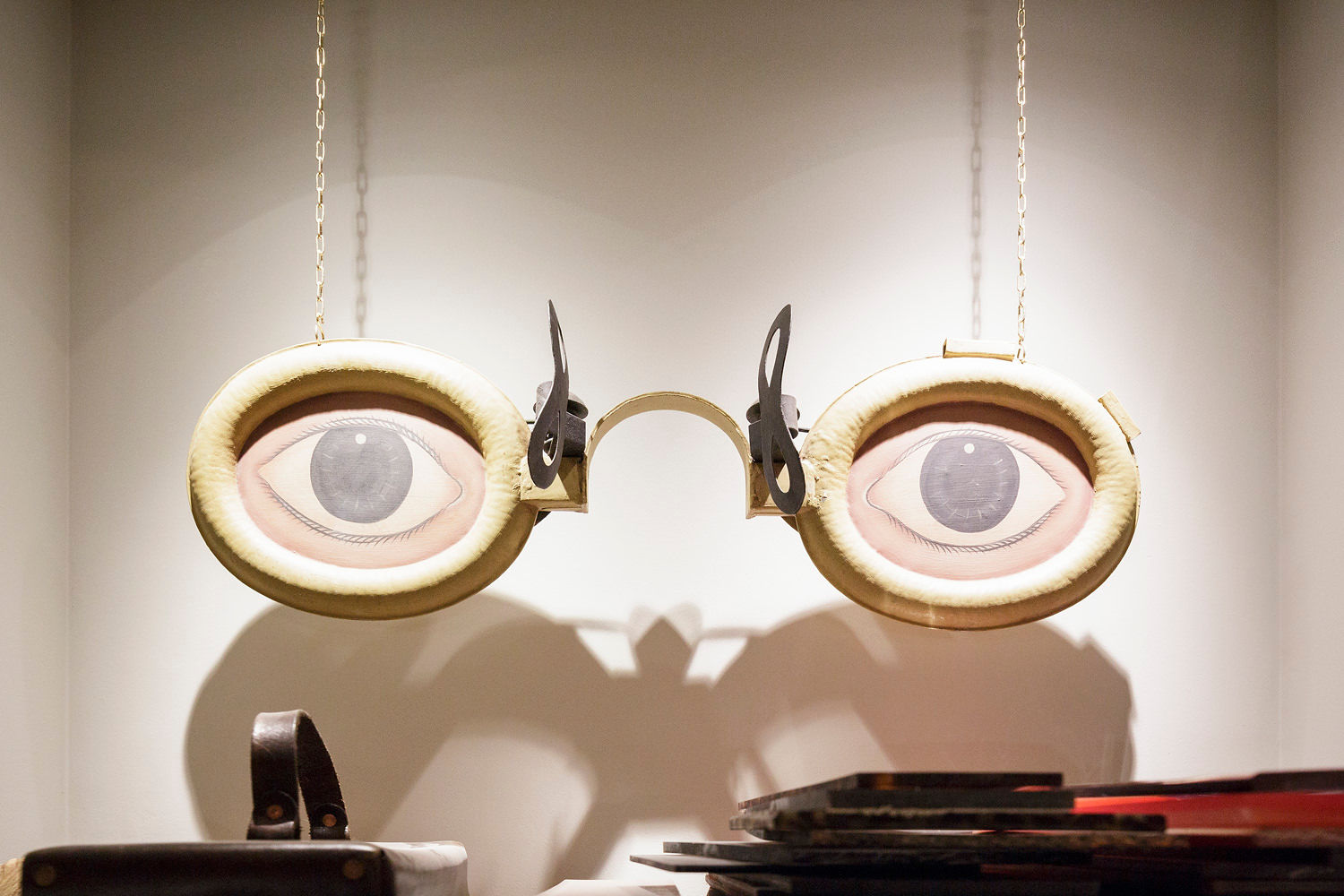 Large, whimsical eyeglasses sculpture with realistic eyes set against a plain background, suspended by chains. Shadowy objects appear below the sculpture, evoking an abstract interpretation of Understanding Posterior Vitreous Detachment.