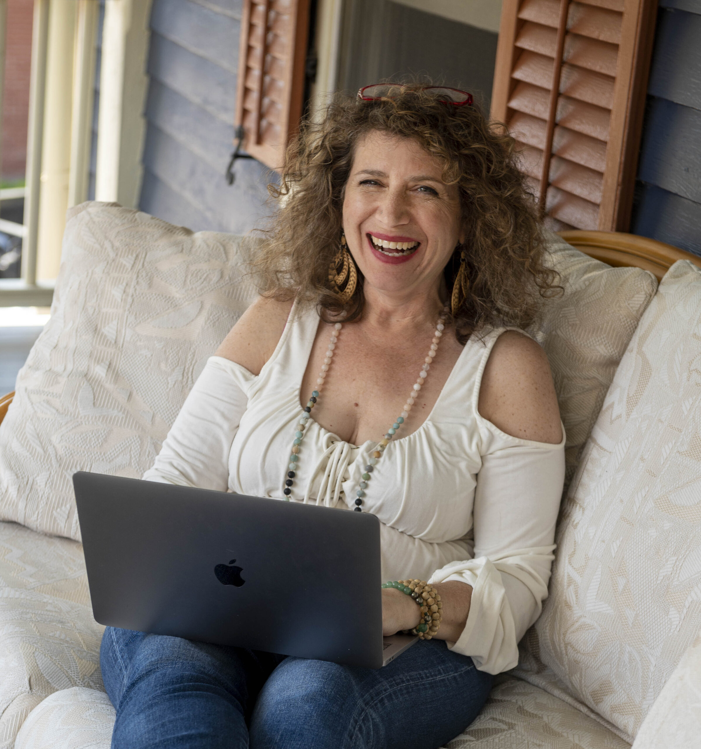 Junie Moon with curly hair wearing a white off-shoulder top and jeans sits on a cushioned bench, smiling while using a laptop. She's browsing Who-is-Junie-Moon, clearly captivated by the fascinating stories.