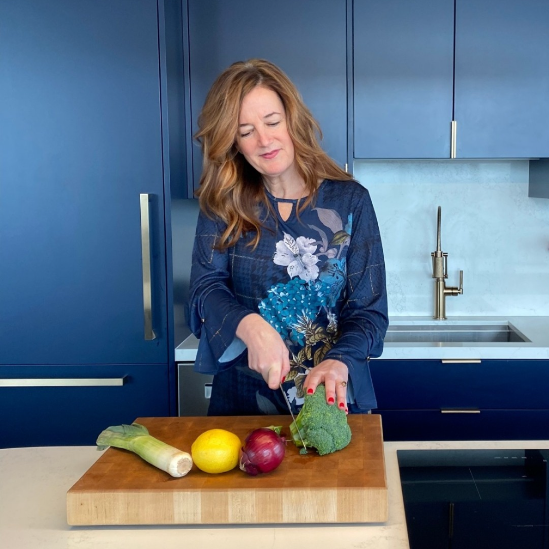 Annie Gaudreault is cutting broccoli on a wooden cutting board in a modern kitchen with blue cabinets. Other vegetables, including a leek, red onion, and lemon, are also on the cutting board, emphasizing eating for vitality: the importance of fiber.