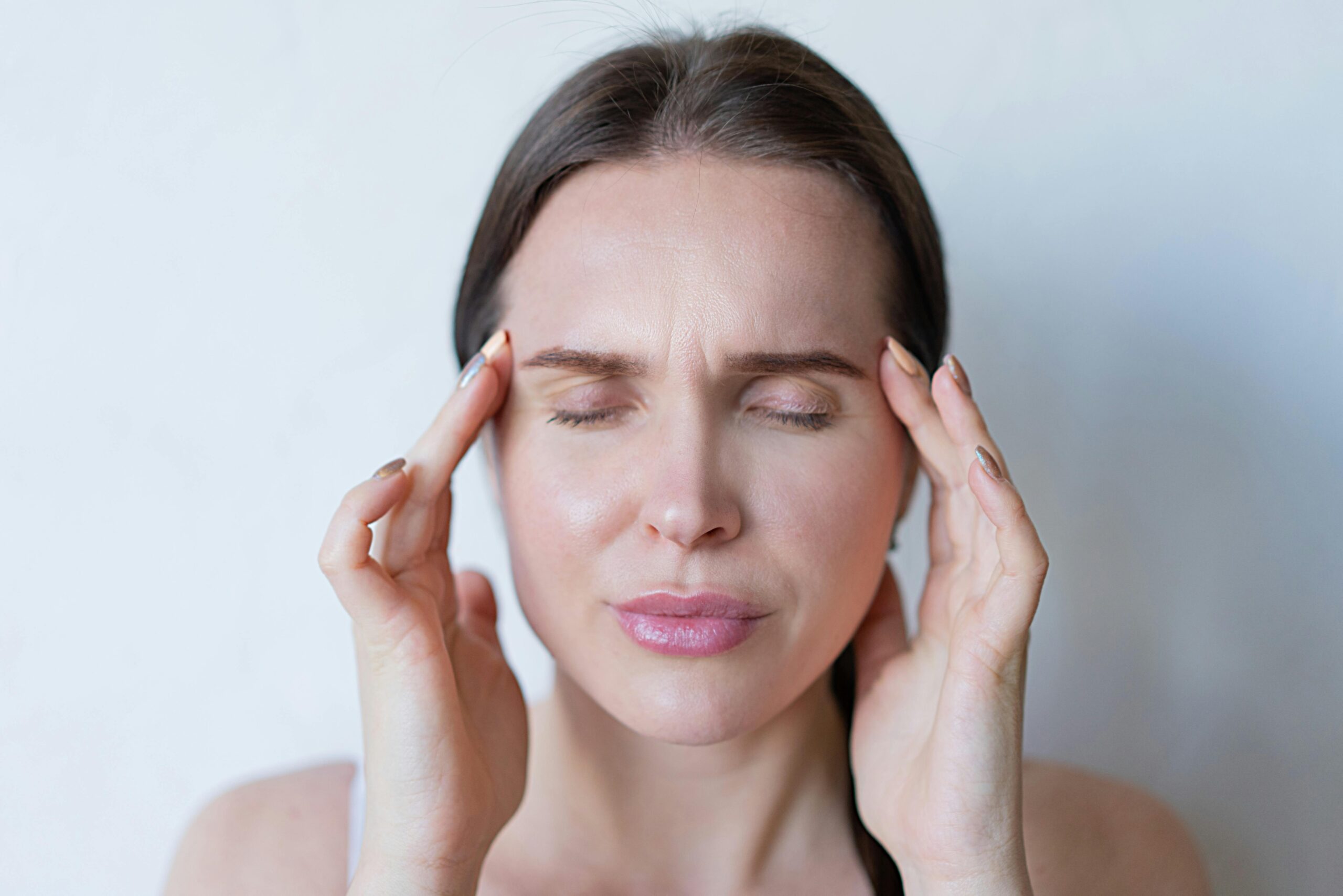 A woman with closed eyes touches her temples with both hands, appearing to be in discomfort or deep thought, as if she is weighing the balancing benefits and risks of  HRT.