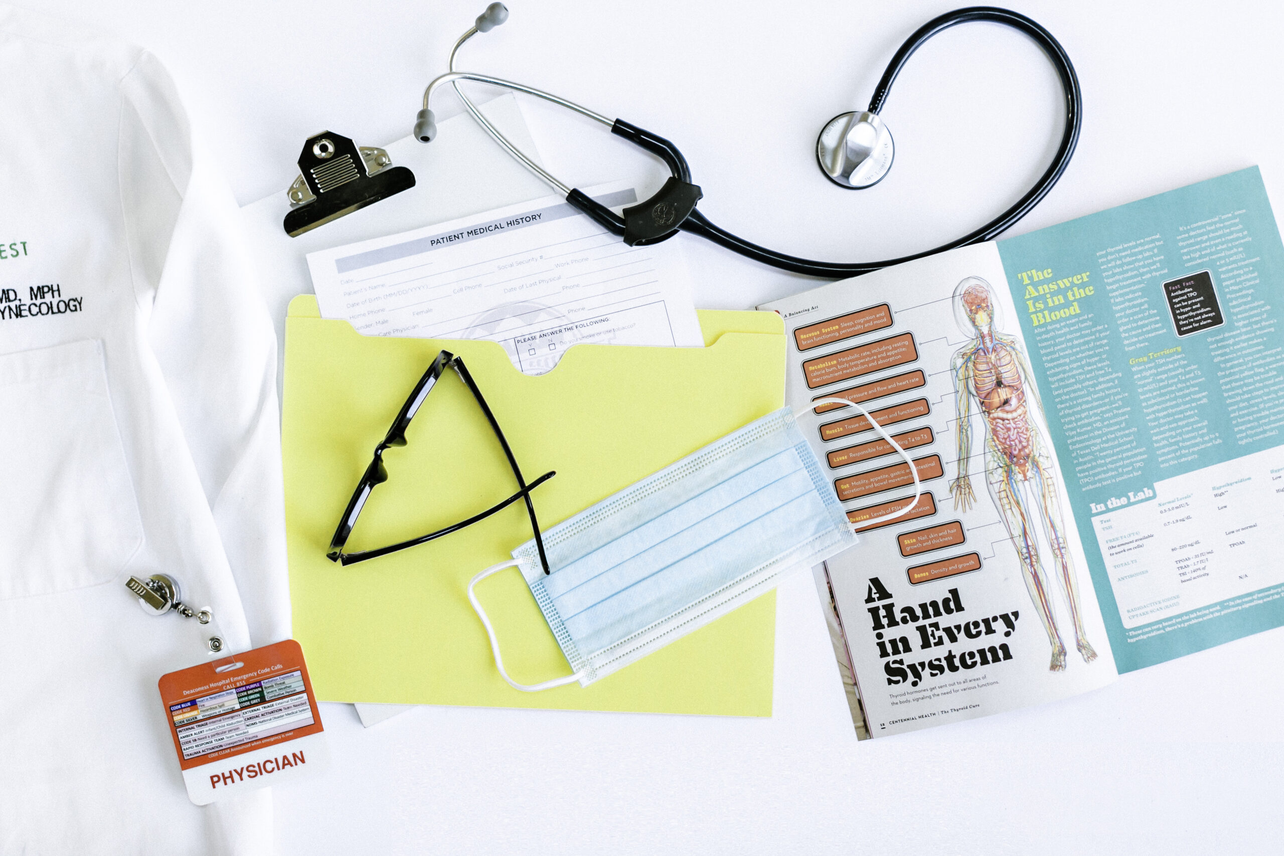 A doctor's lab coat, stethoscope, clipboard with paperwork detailing postmenopausal HRT, surgical mask, glasses, yellow folder marked "90 Day Journey," and anatomical charts on a white surface.