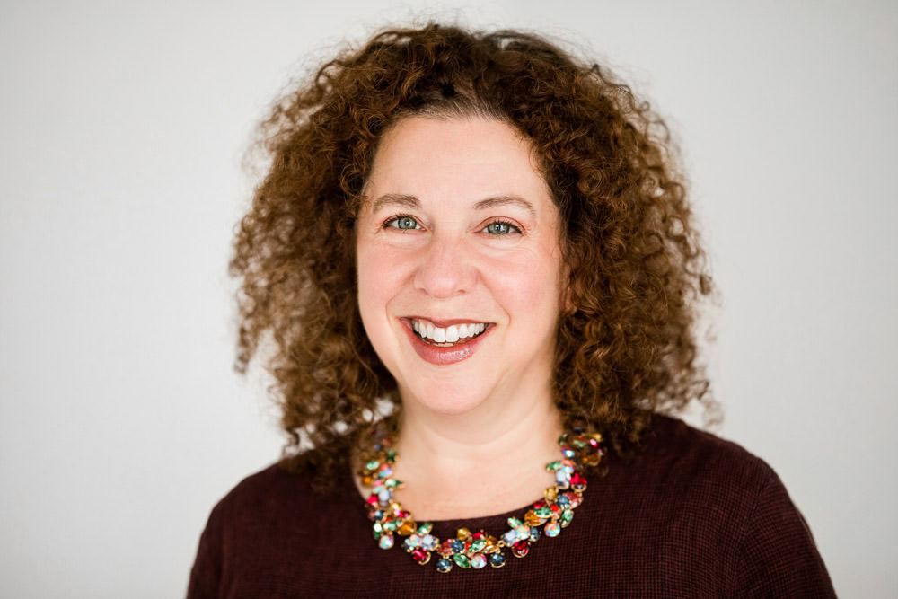 A woman with curly brown hair is smiling at the camera, wearing a colorful beaded necklace and a dark top. She is one of our exciting upcoming guests.