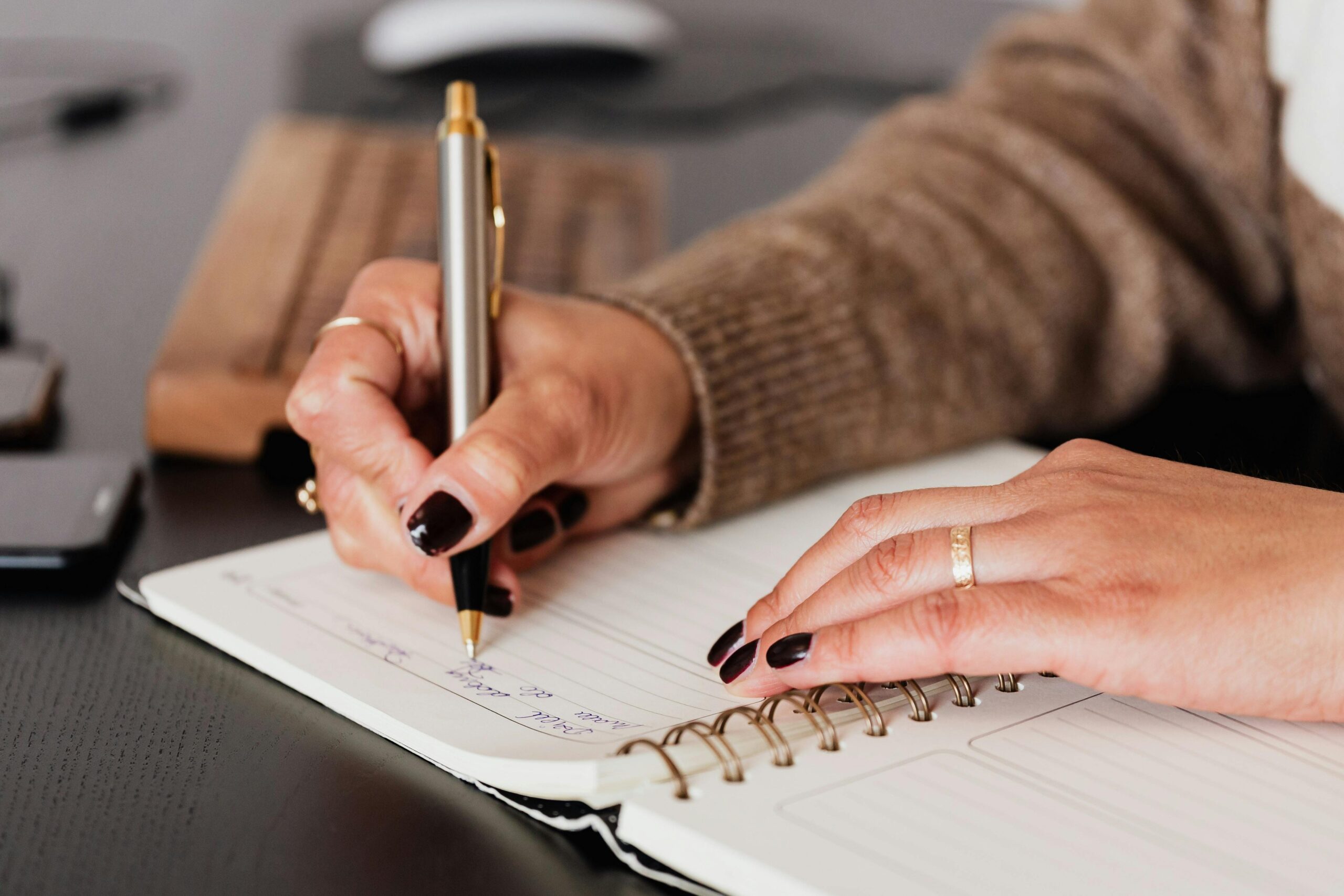 A person with dark nail polish writes in a spiral-bound notebook with a pen on a desk containing a wooden keyboard and a smartphone, possibly jotting down notes about their experience with Hormone Replacement Therapy.