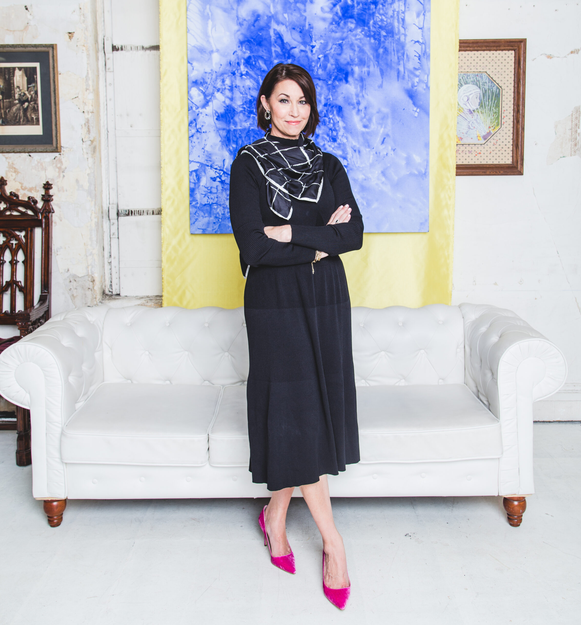 Meet Alison Bruhn: A person stands confidently with arms crossed in a room with white walls. Dressed in a black outfit and pink shoes, she stands before a white sofa, with a blue painting adorning the wall behind her.