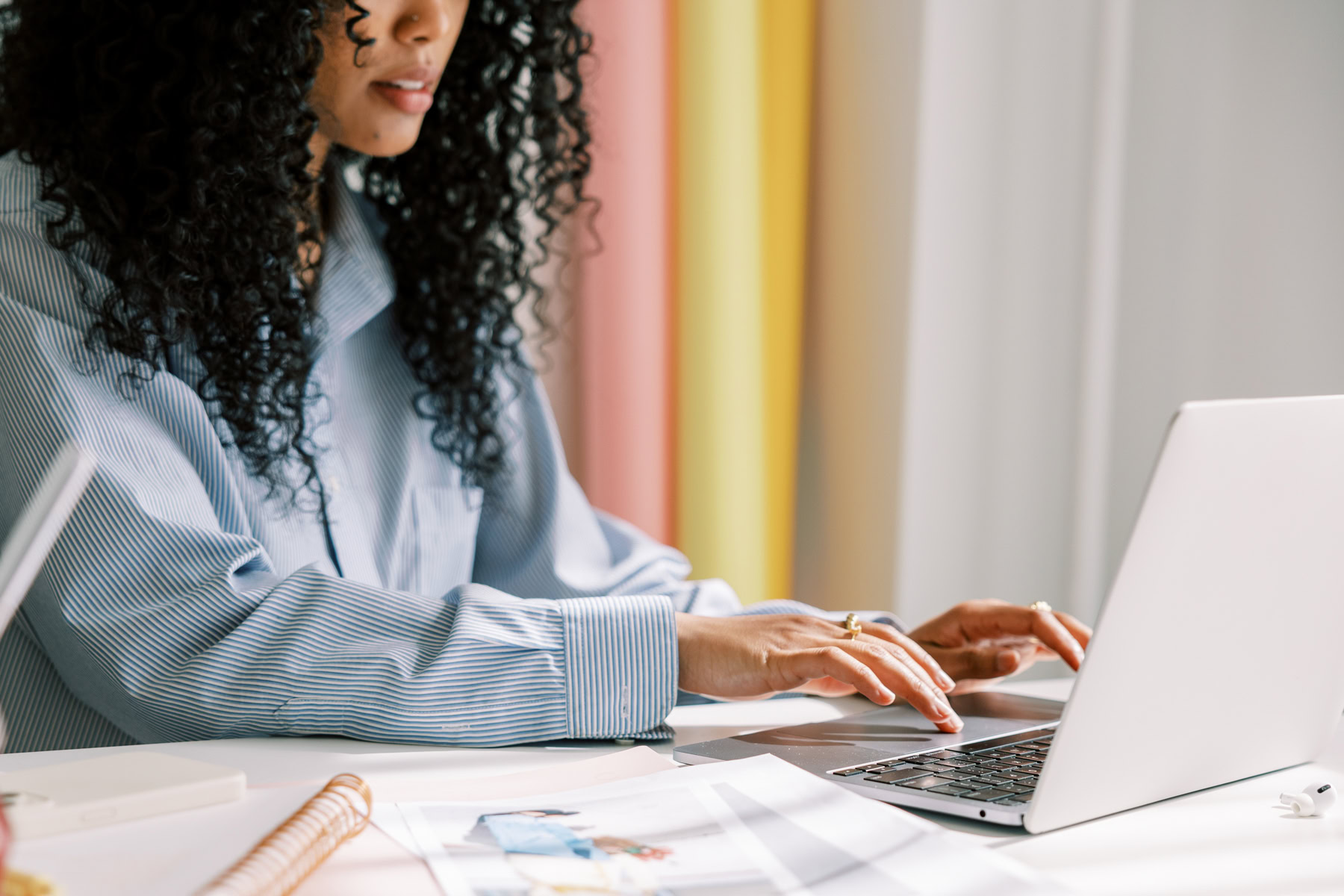 Person with curly hair typing on a laptop at a desk, surrounded by various papers and documents discussing postmenopausal HRT, evaluating the risks and potential relief it offers.