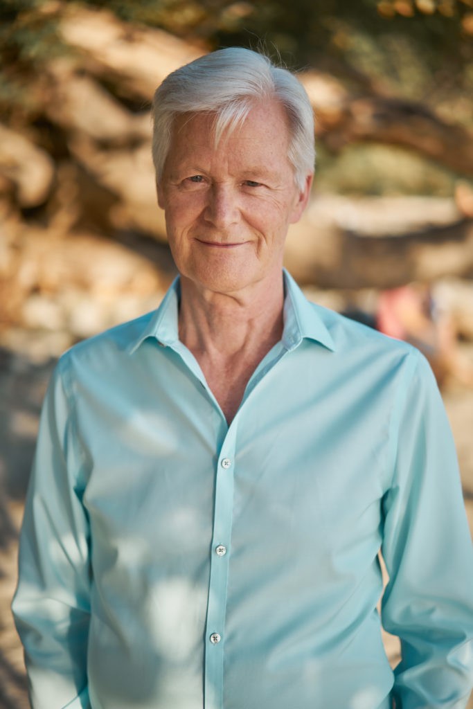 An older man with white hair, wearing a light blue button-up shirt, smiles at the camera. As our upcoming guest, his presence promises to add a unique touch to the event focused on empowerment. The background shows a blurry outdoor setting.