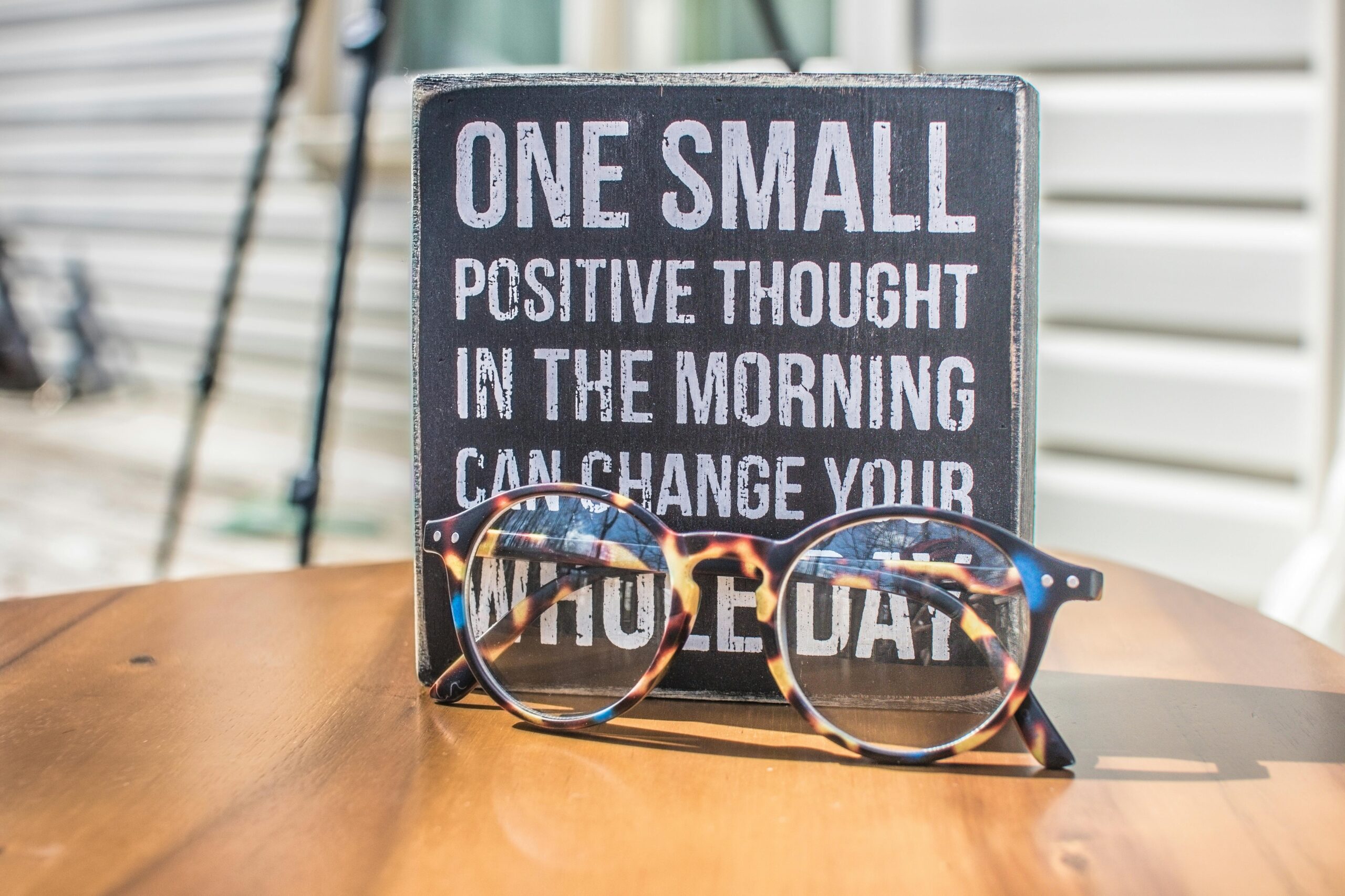 A plaque with the text "One small positive thought in the morning can change your whole day" is placed on a wooden surface next to a pair of tortoiseshell glasses, serving as a gentle reminder of the importance of understanding negative thoughts and their impact.