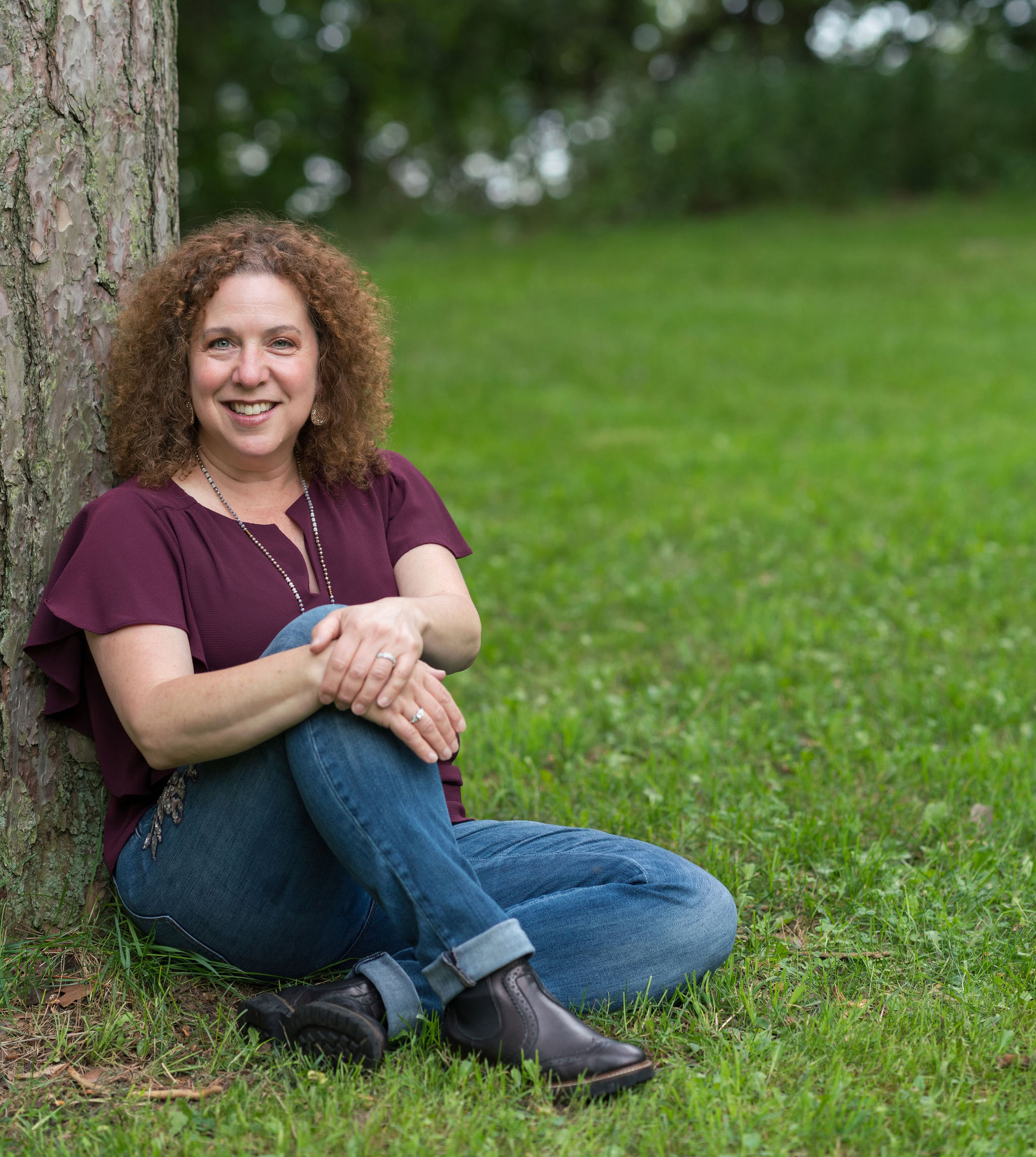 Against the sturdy trunk of a tree, Lynne Golodner sits on green grass, smiling warmly. Her curly hair cascades over a burgundy top paired with blue jeans and black boots, embodying the essence of building a writing community for support and growth in her surroundings.