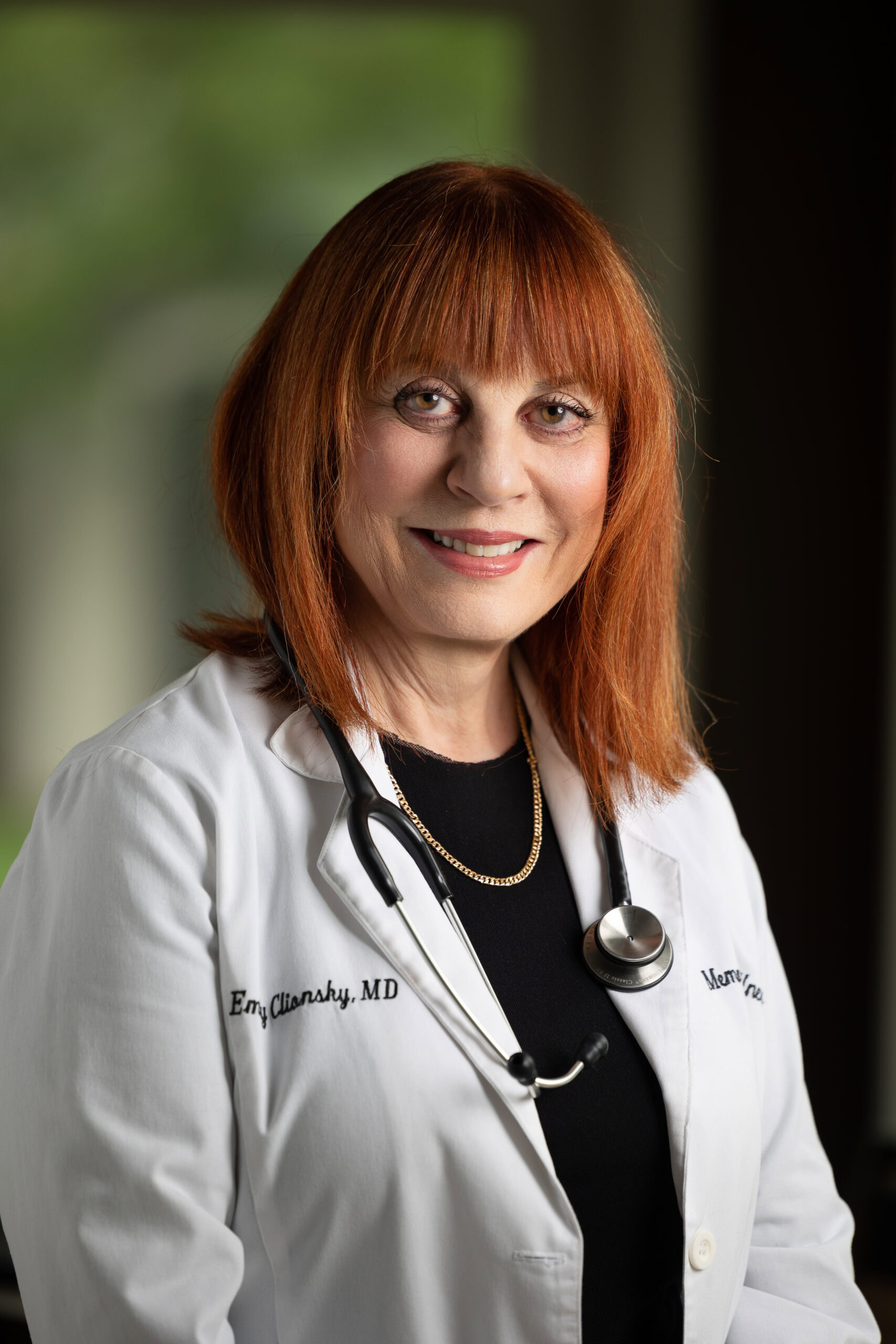 Dr. Emily Clionsky with red hair wearing a white lab coat and a stethoscope around their neck stands indoors, focusing on nutrition's role in preventing cognitive decline.