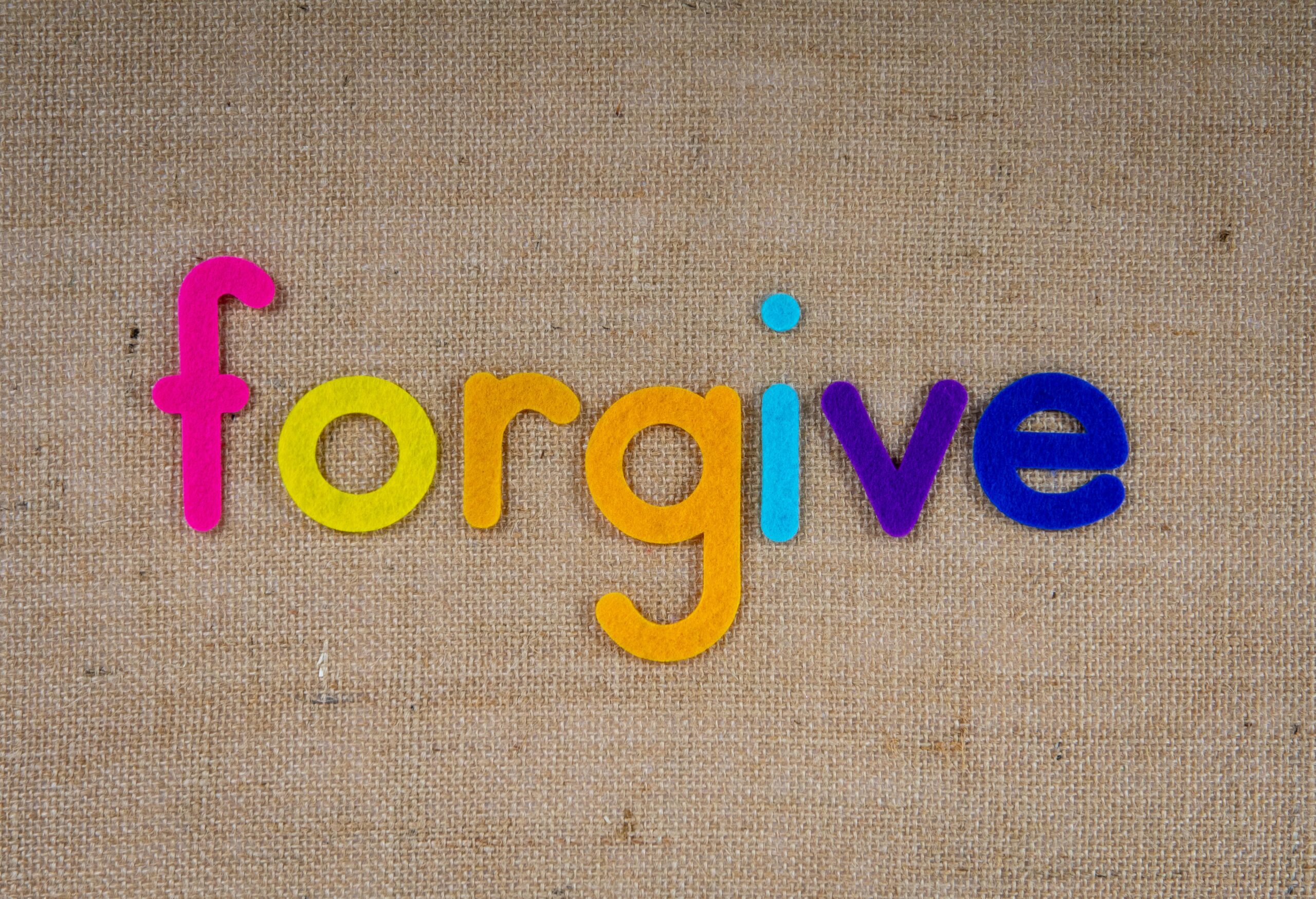 Colorful felt letters spell "forgive" on a burlap background, serving as a gentle reminder to release energy drainers and stop holding grudges.