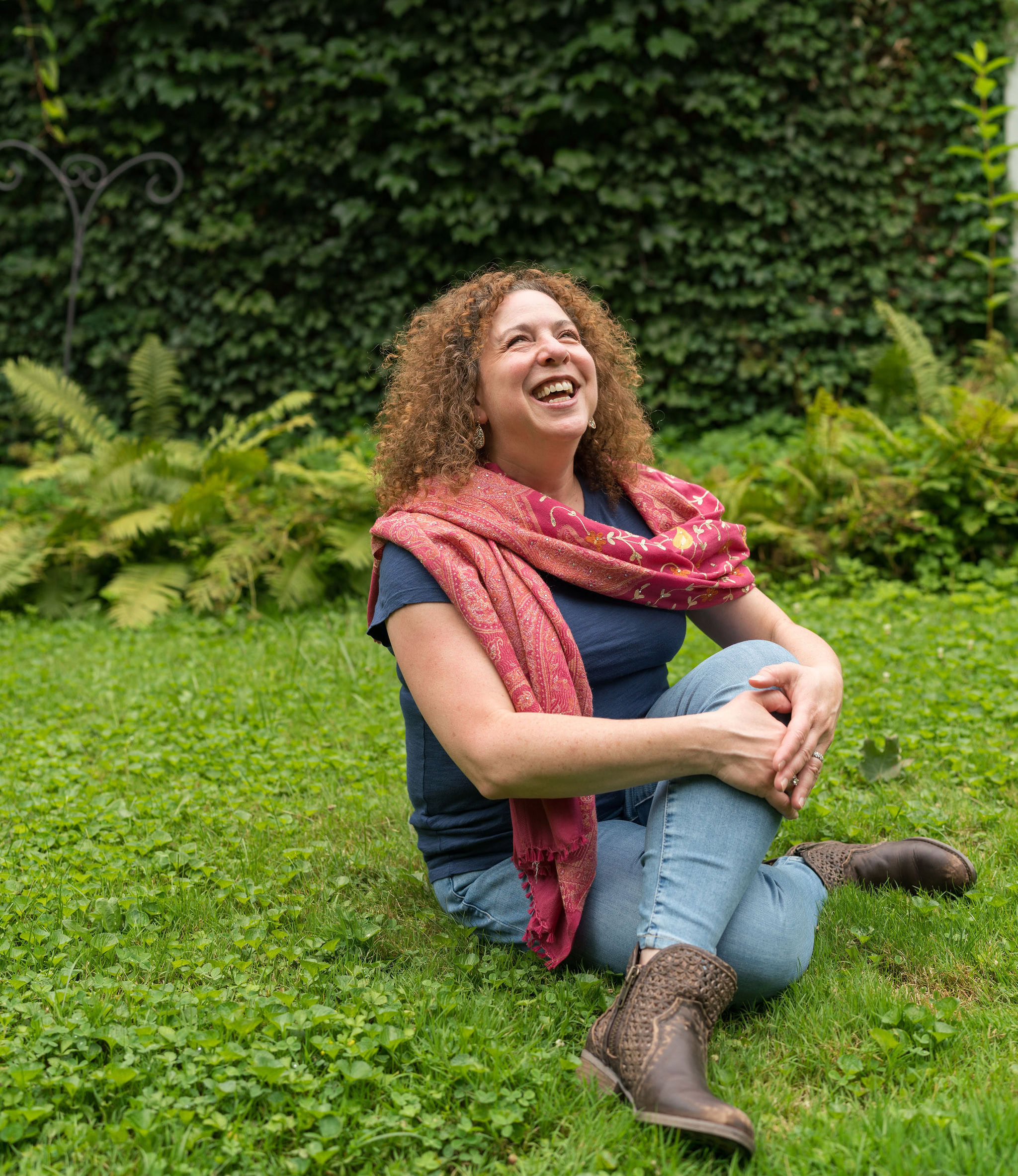 Amid the challenges, Lynne Golodner with curly hair sits on the grass, finding inspiration as they smile and look up. Dressed in jeans, a blue shirt, and a patterned pink scarf, they are surrounded by ferns and an ivy-covered wall that offers structure to the scene.