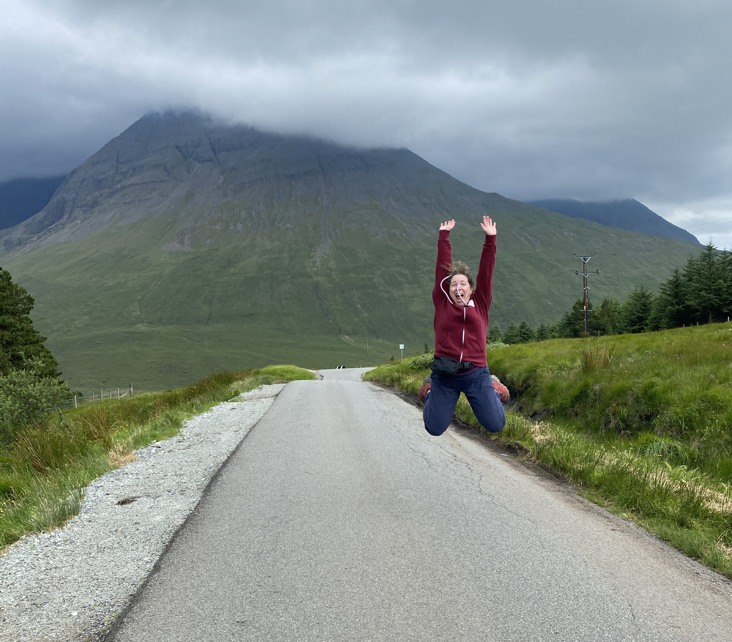 Amidst a mountainous landscape under a cloudy sky, Lynne Golodner in a red hoodie leaps onto the road, embodying the liberating spirit of "The Journey of Writing as Therapy.