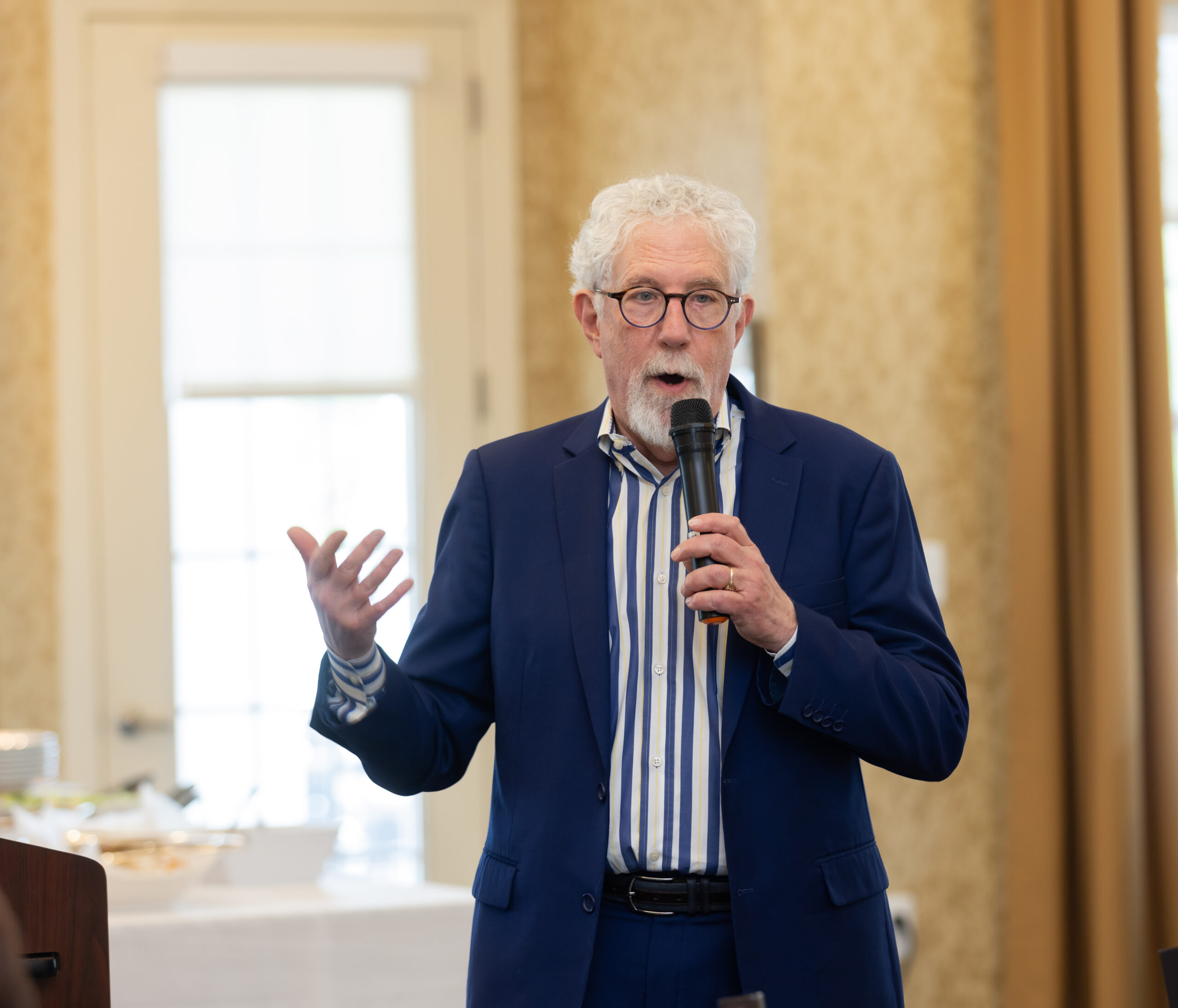 Dr. Mitchell Clionsky with white hair and a beard is speaking into a microphone about social connections, gesturing with his hand. He is wearing glasses, a blue suit, and a striped shirt.