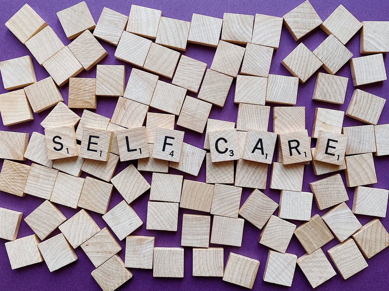 Wooden letter tiles spelling "SELF CARE" are surrounded by blank tiles on a purple background, subtly hinting at overcoming self-sabotage.