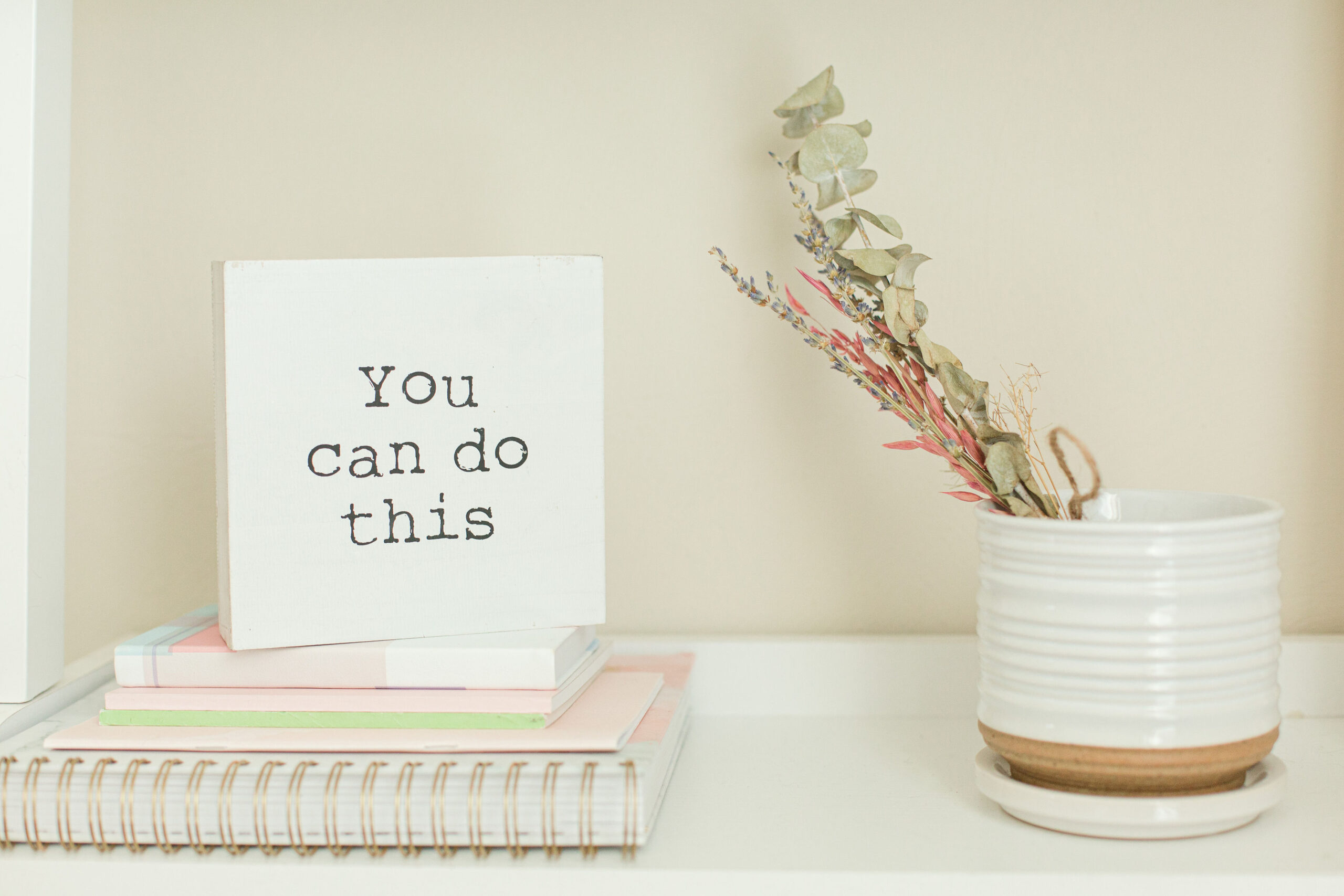 A small sign with "You can do this" offers practical tips to boost your energy today, nestled among stacked books and a white mug with dried flowers on a shelf.