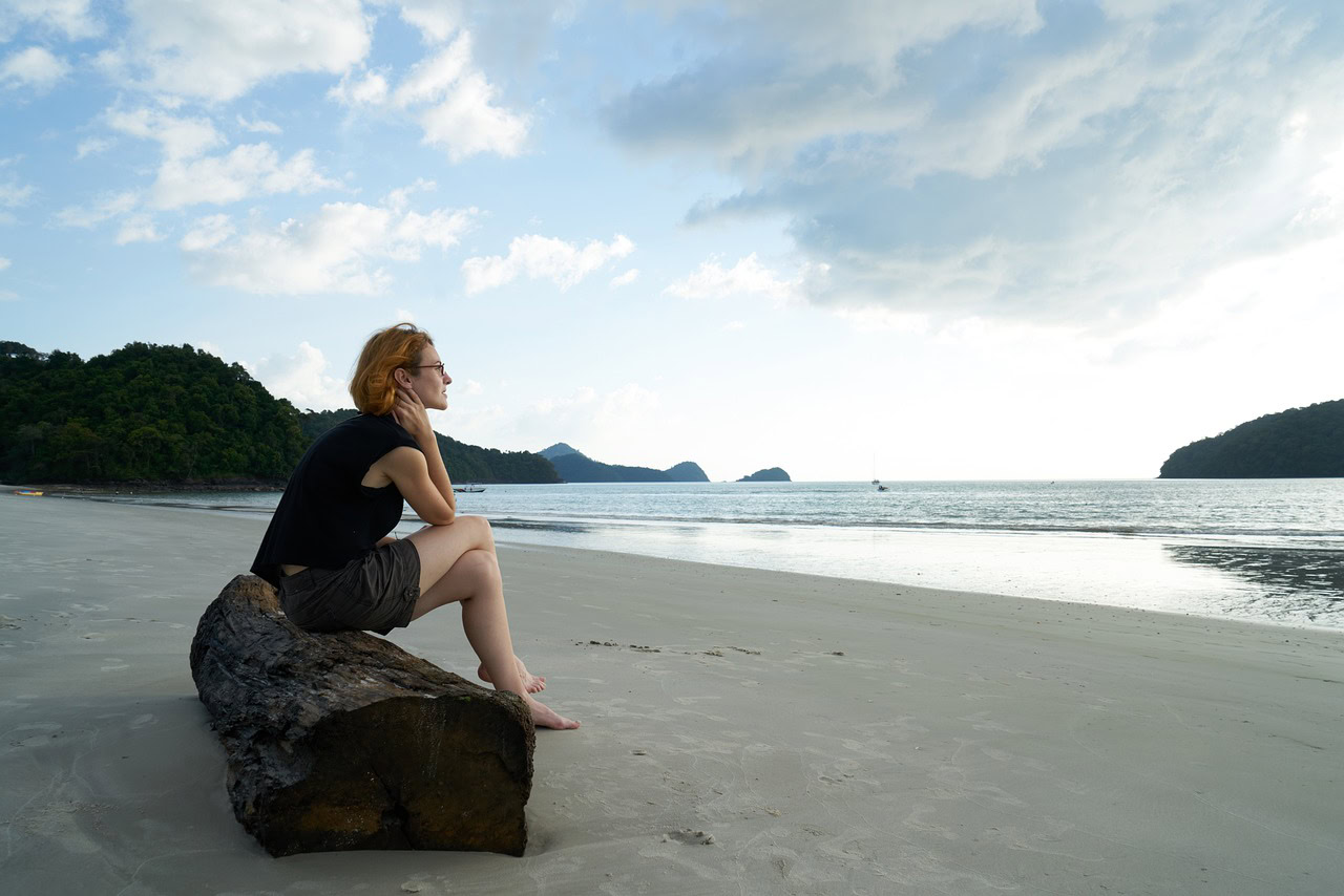 A person sits on a log at the beach, contemplating the sea under a cloudy sky. Mountains loom and a boat drifts in the distance, as if shedding stress and leaving behind energy drainers with each gentle wave.