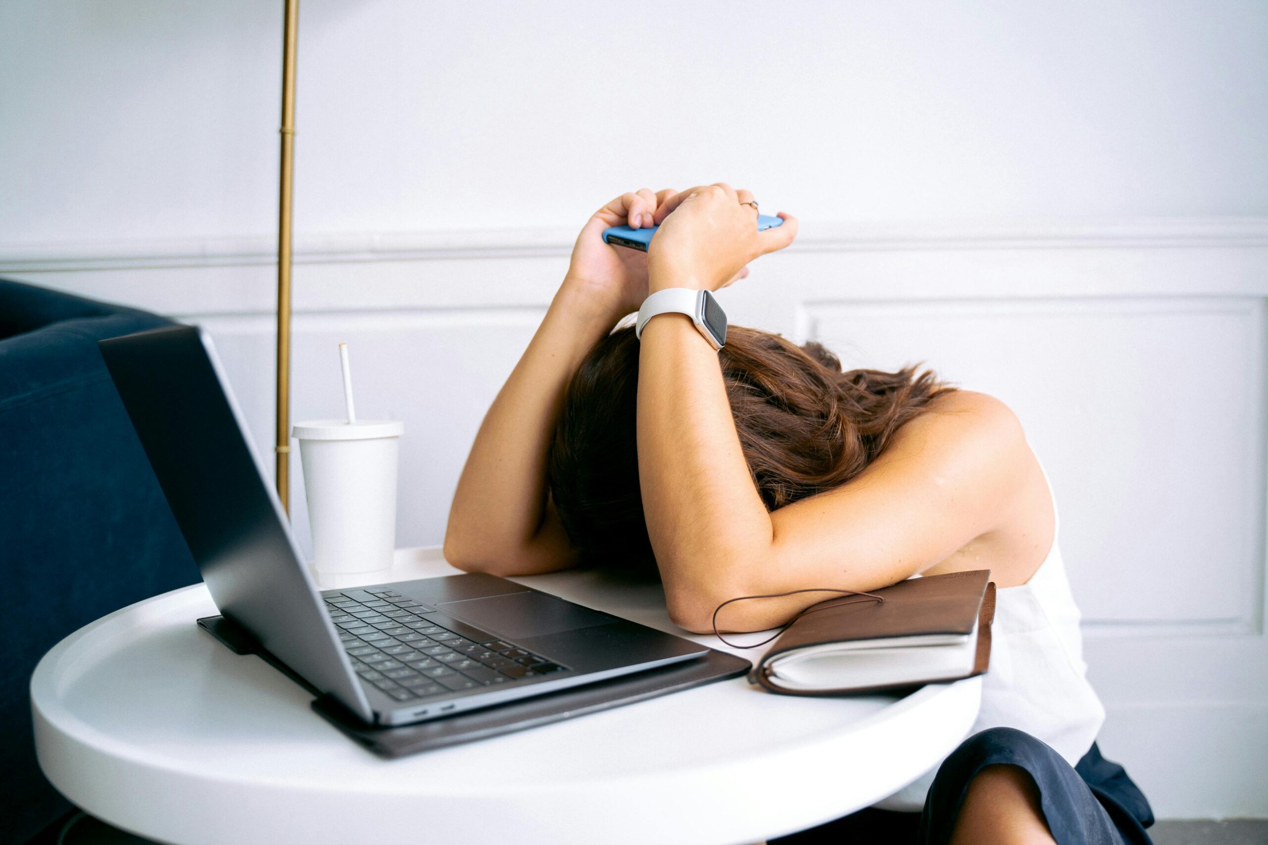 Person sitting at a table with their head down, holding a phone, surrounded by a laptop and a notebook, deep in thought as they reflect on understanding self-sabotage and how it impacts their daily productivity.