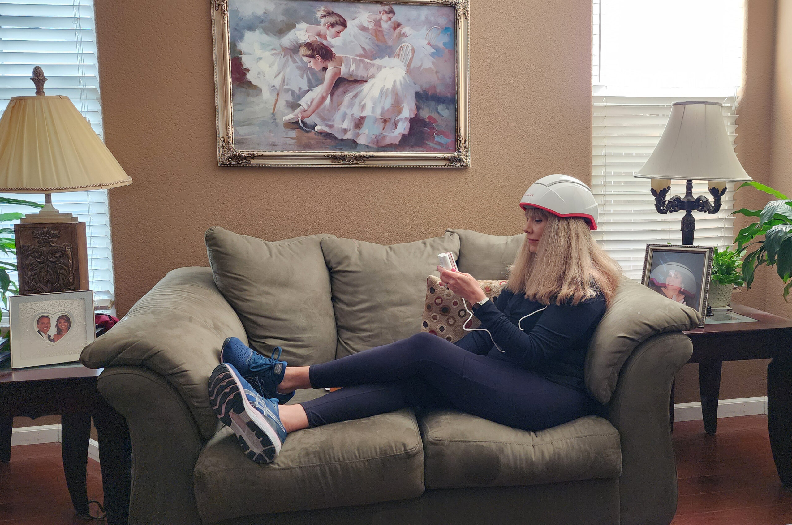 Carmen Hecox sits on a couch wearing a white helmet for low-level light therapy for hair growth, holding power controller. A painting of ballerinas graces the wall, with a lamp casting a warm glow on each side table.