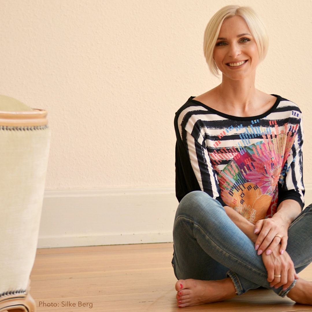 Anne-Sophie Reinhardt with short blonde hair sits confidently on a wooden floor, wearing a colorful top and jeans, smiling radiantly at the camera, embodying body positivity.