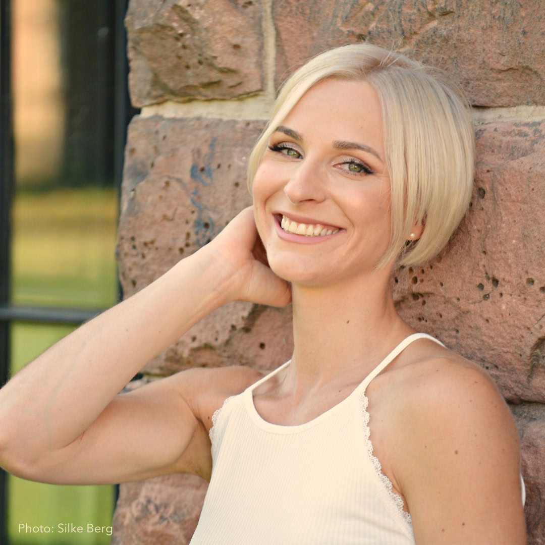 Anne-Sophie Reinhardt with short blonde hair smiles confidently while leaning against a brick wall, embracing self-acceptance in her light-colored tank top.