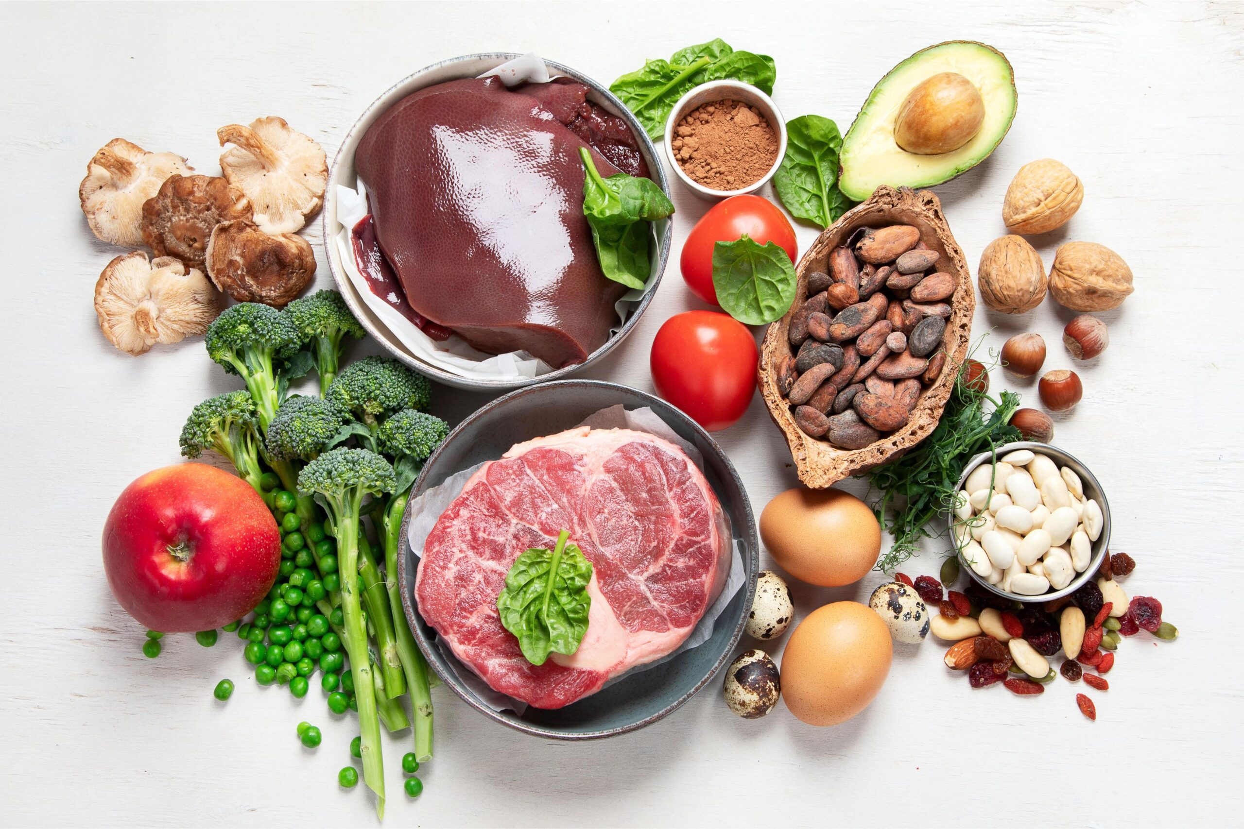 A variety of iron-rich foods, ideal for increasing iron and ferritin levels safely, includes raw meat, chicken liver, broccoli, nuts, beans, eggs, avocado, tomatoes, peas, mushrooms, an apple, and cocoa.