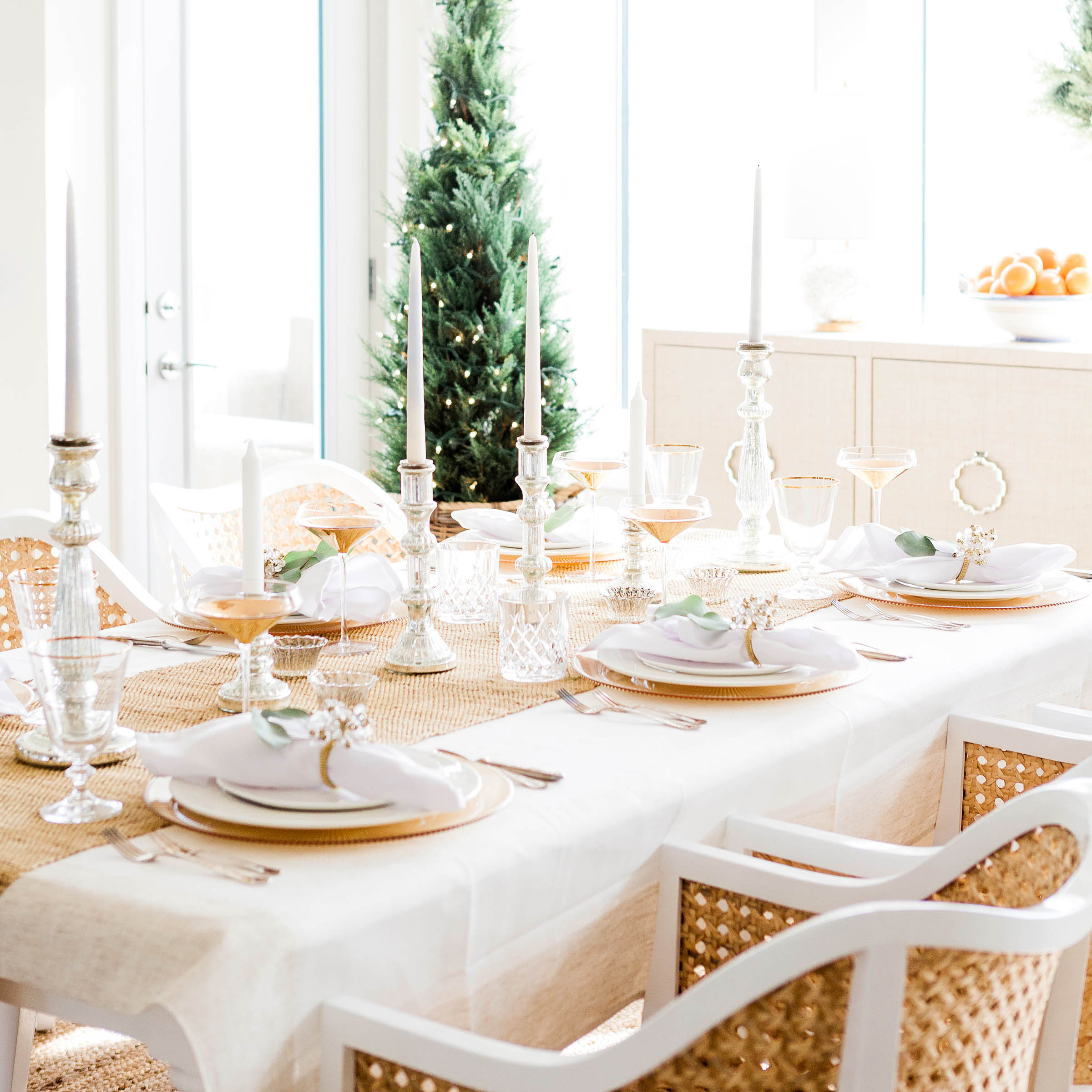 A festive dining table is elegantly set with white plates, crystal glassware, and silver candlesticks, exuding a calm sophistication. A Christmas tree stands gracefully in the background, adding to the serene holiday ambiance.