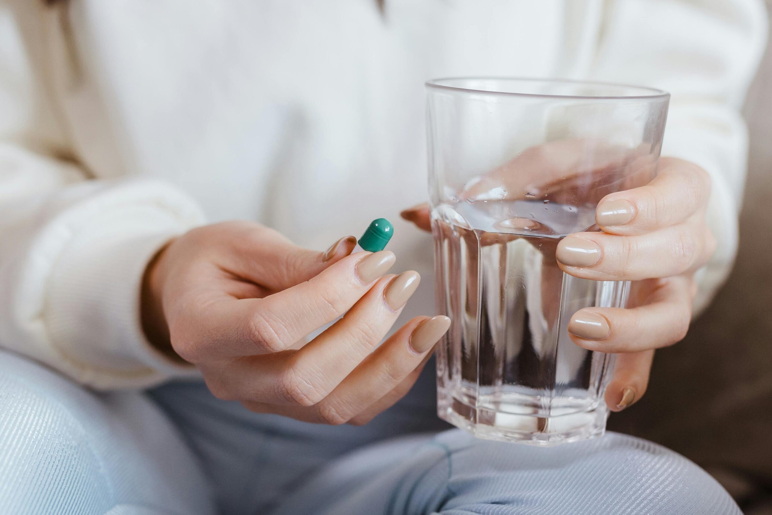 A person holds a green capsule in one hand and a glass of water in the other, hoping to nourish their body with essential nutrients like iron, which is vital for preventing hair loss.