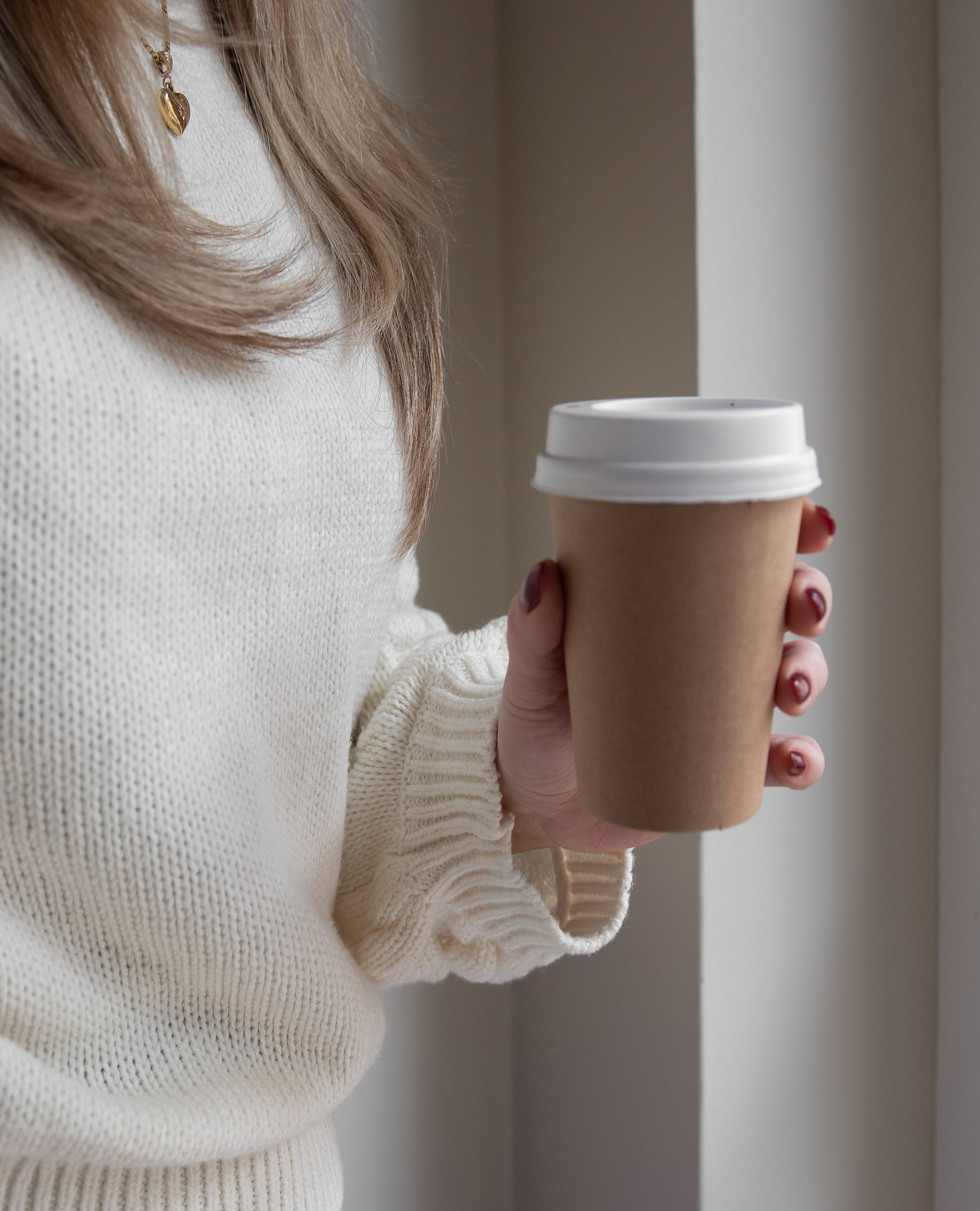 Person in a white sweater holding a brown disposable coffee cup with a white lid near a window, savoring the moment and practicing mindfulness to find holiday peace.