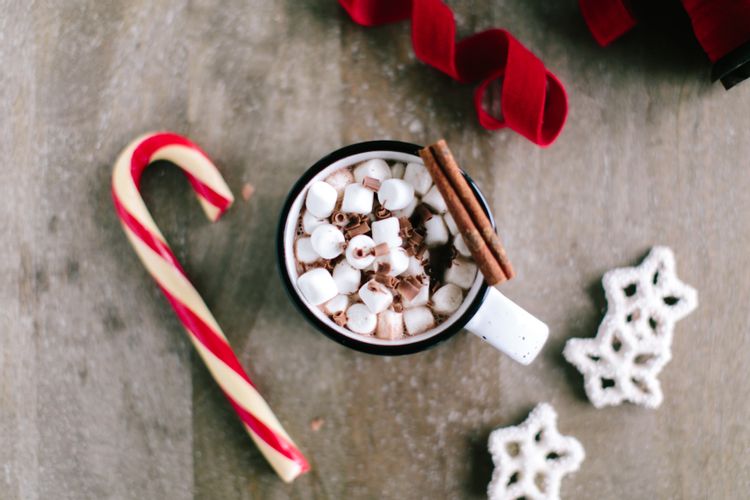 A cup of hot chocolate topped with marshmallows and a cinnamon stick sits calmly on a table, surrounded by a candy cane and decorative white snowflakes, creating the perfect serene holiday scene.