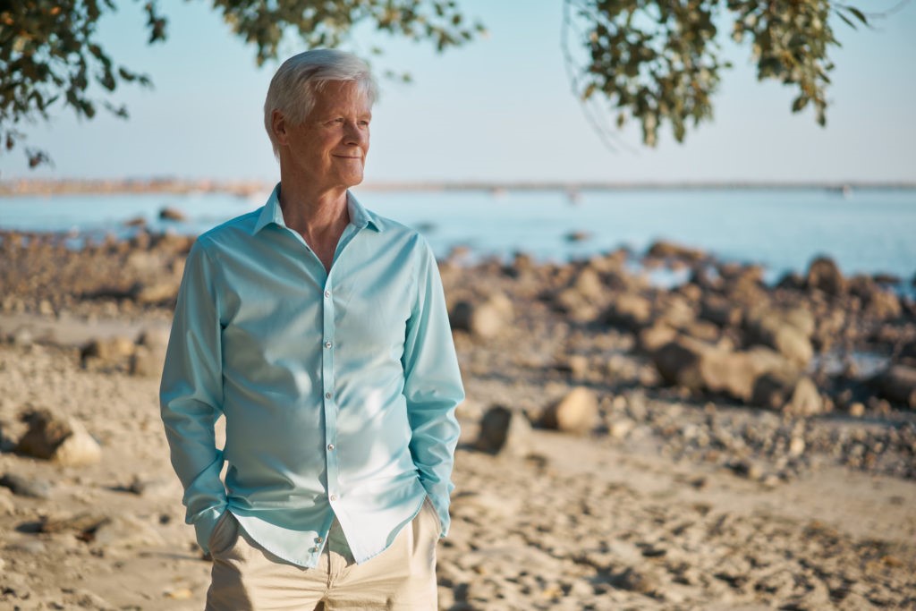 Udo Erasmus in a light blue shirt stands on a rocky beach, hands in his pockets, contemplating the horizon as he reflects on exploring Udo's personal health journey.