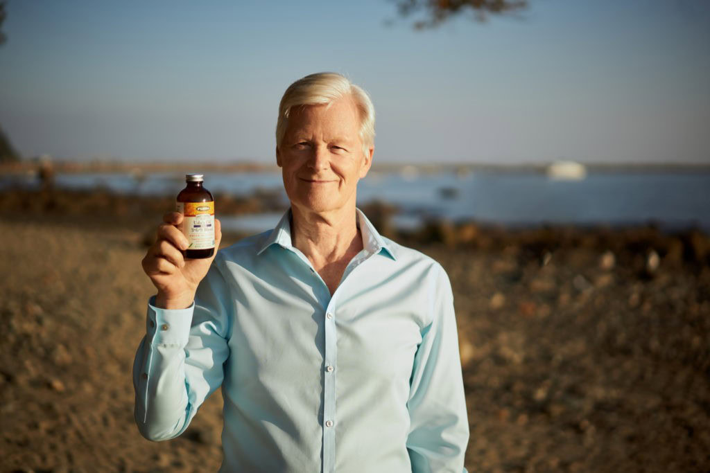 Udo Erasmus in a light blue shirt holds a small bottle of healthy oils while standing on a beach with the sea in the background, embracing midlife wellness.