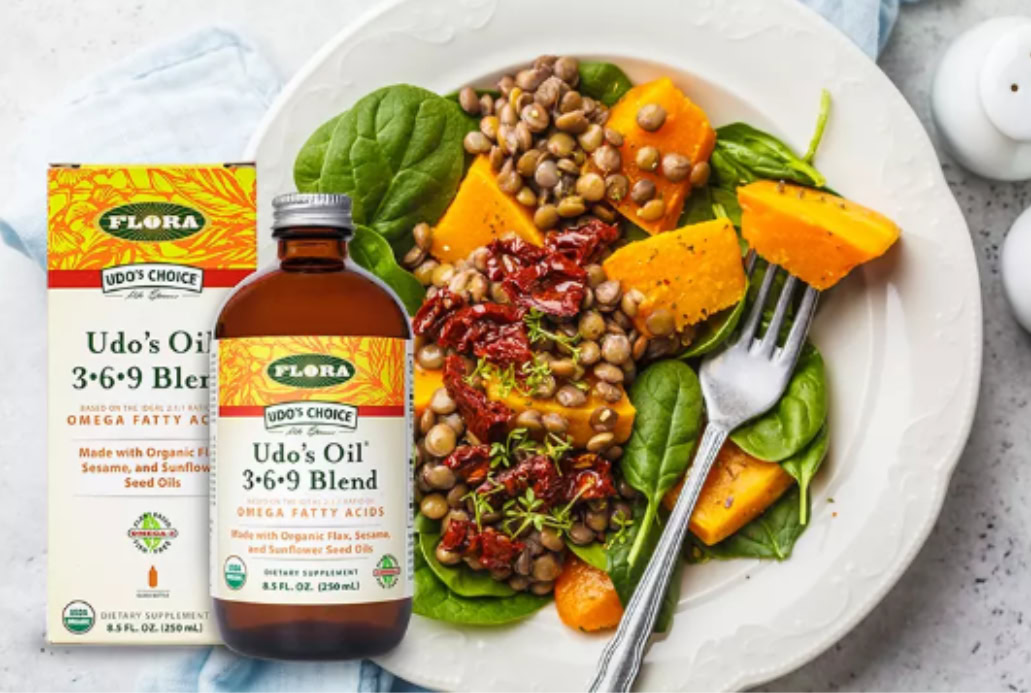 A plate of spinach, sweet potato, lentils, and sun-dried tomatoes sits beside a bottle and box of Udo's Oil 3•6•9 Blend, a perfect addition for those embracing nutrition and midlife wellness.