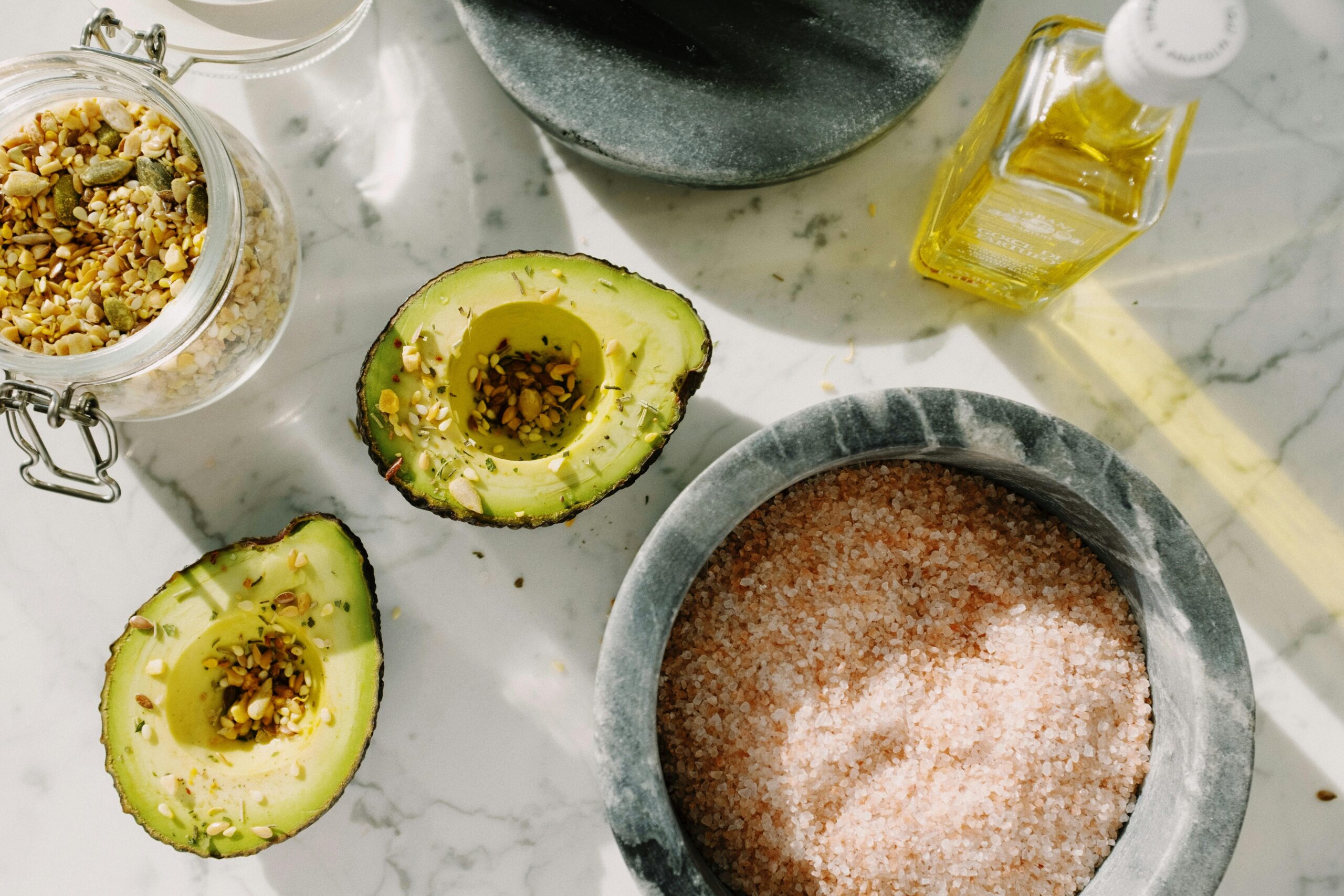 A halved avocado sits on a marble counter, rich in Omega 3 and 6 essential for women's brain and body health, next to a jar of grains, a bottle of oil, and a bowl of pink salt.