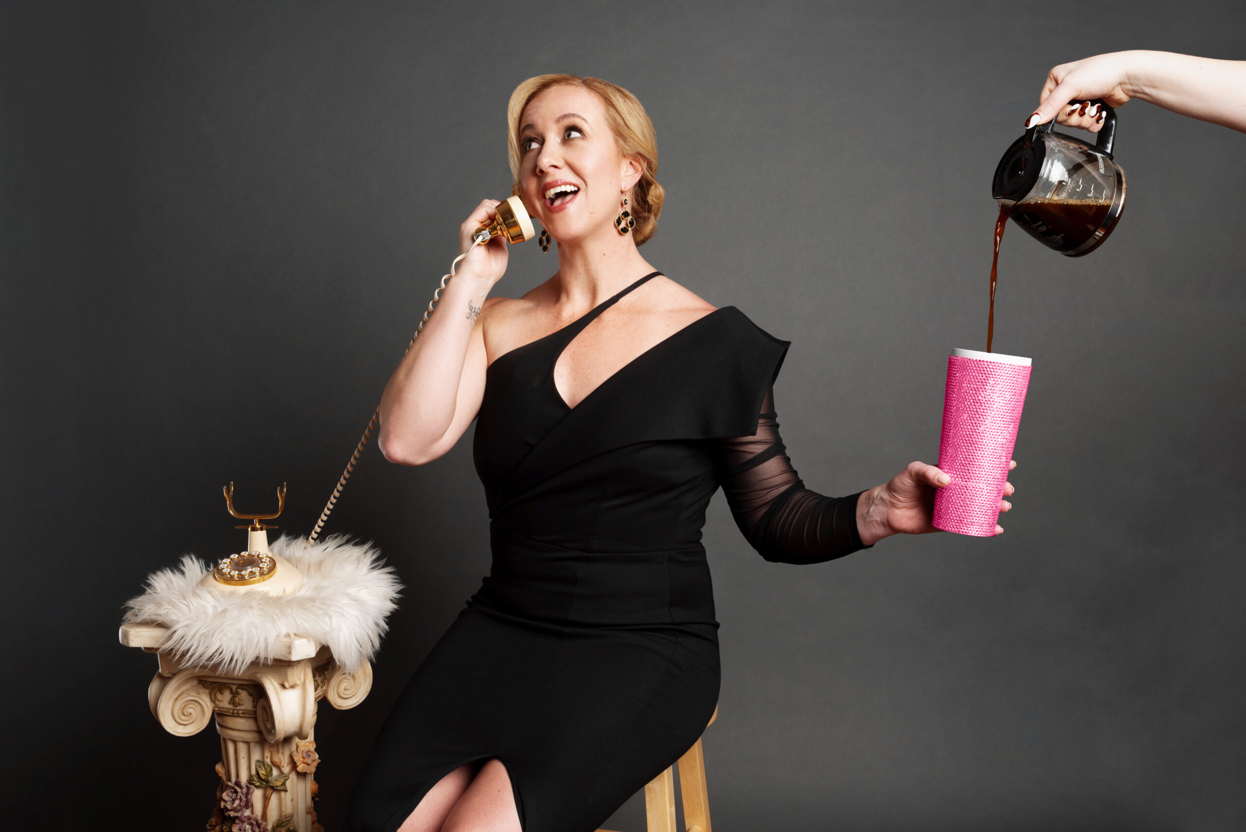 A woman in a black dress, embodying the power of saying yes, talks on a retro phone while holding a pink cup. As coffee is poured into the cup, she sits confidently on a stool beside a small column adorned with a fluffy cloth and phone base, embracing midlife with authenticity and style.