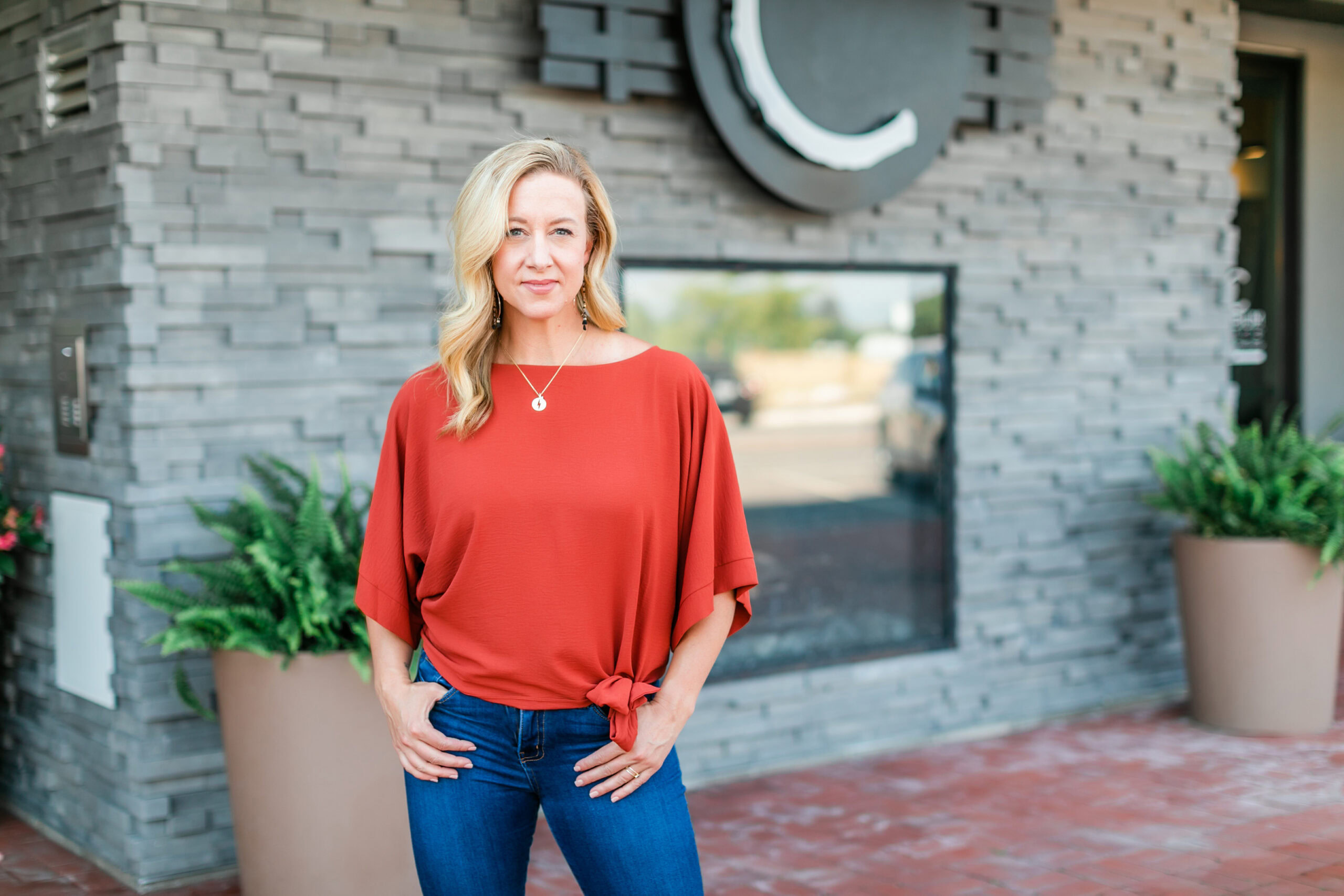 A person stands powerfully in front of a modern brick building, wearing a red blouse and jeans, with hands in pockets. Embracing midlife with confidence, they project a sense of saying "Yes to Yourself.