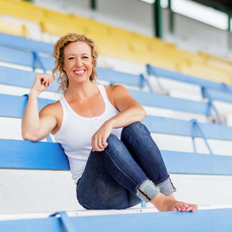 A smiling woman in a white tank top and jeans sits on the bleachers, leaning back casually with one knee up, embodying the confidence of embracing yourself in midlife.