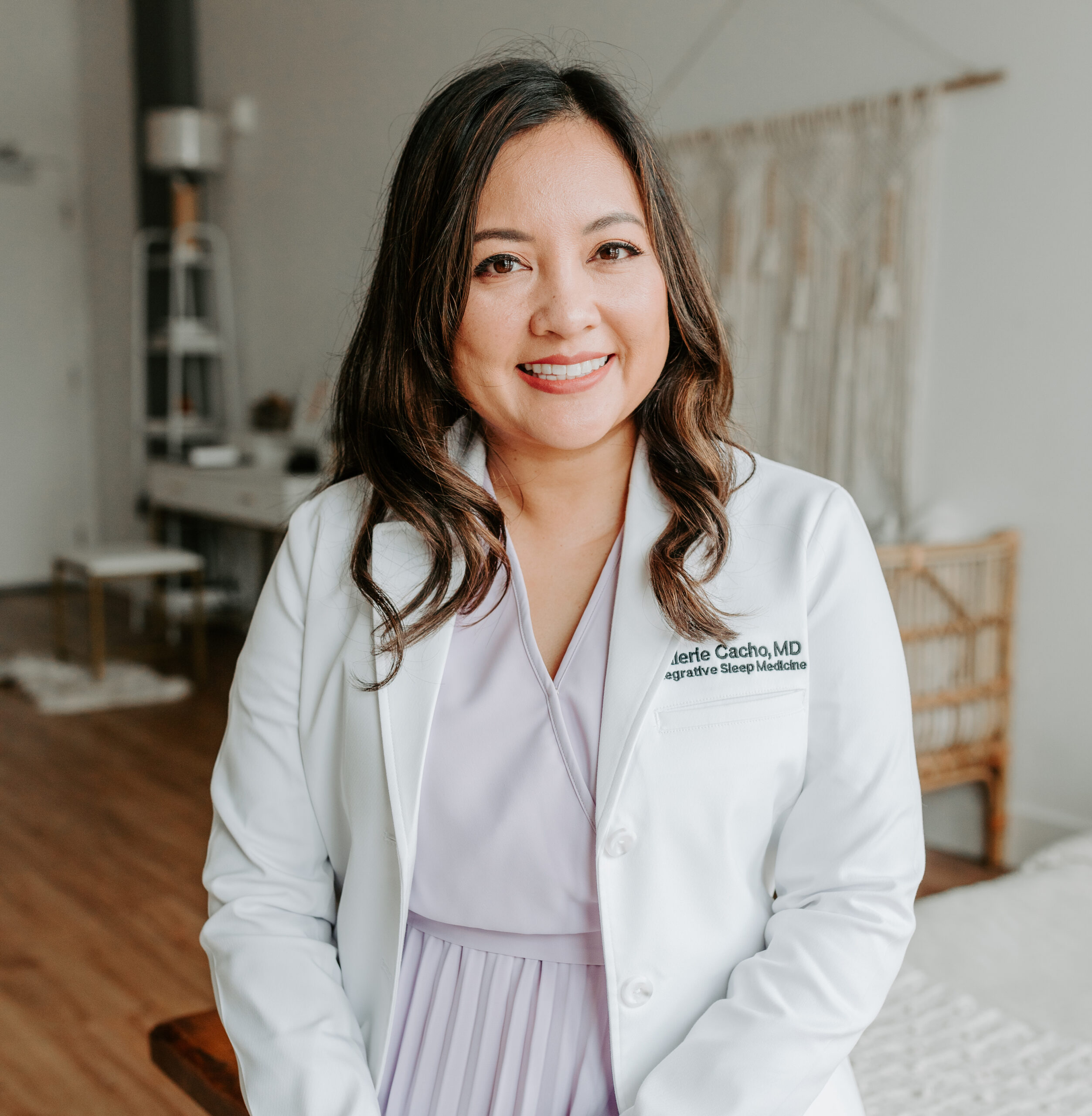 Dr. Val Cacho stands indoors, smiling warmly in her white coat, with medical office furniture behind her. Her lavender dress complements the professional yet welcoming ambiance of the room.