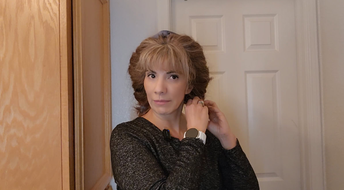 Person standing in front of a door, adjusting hair with a touch of confidence, wearing a long-sleeve dark shirt and a watch. Her heatless curls give an impression of fuller hair, emphasizing the appearance of healthier hair.