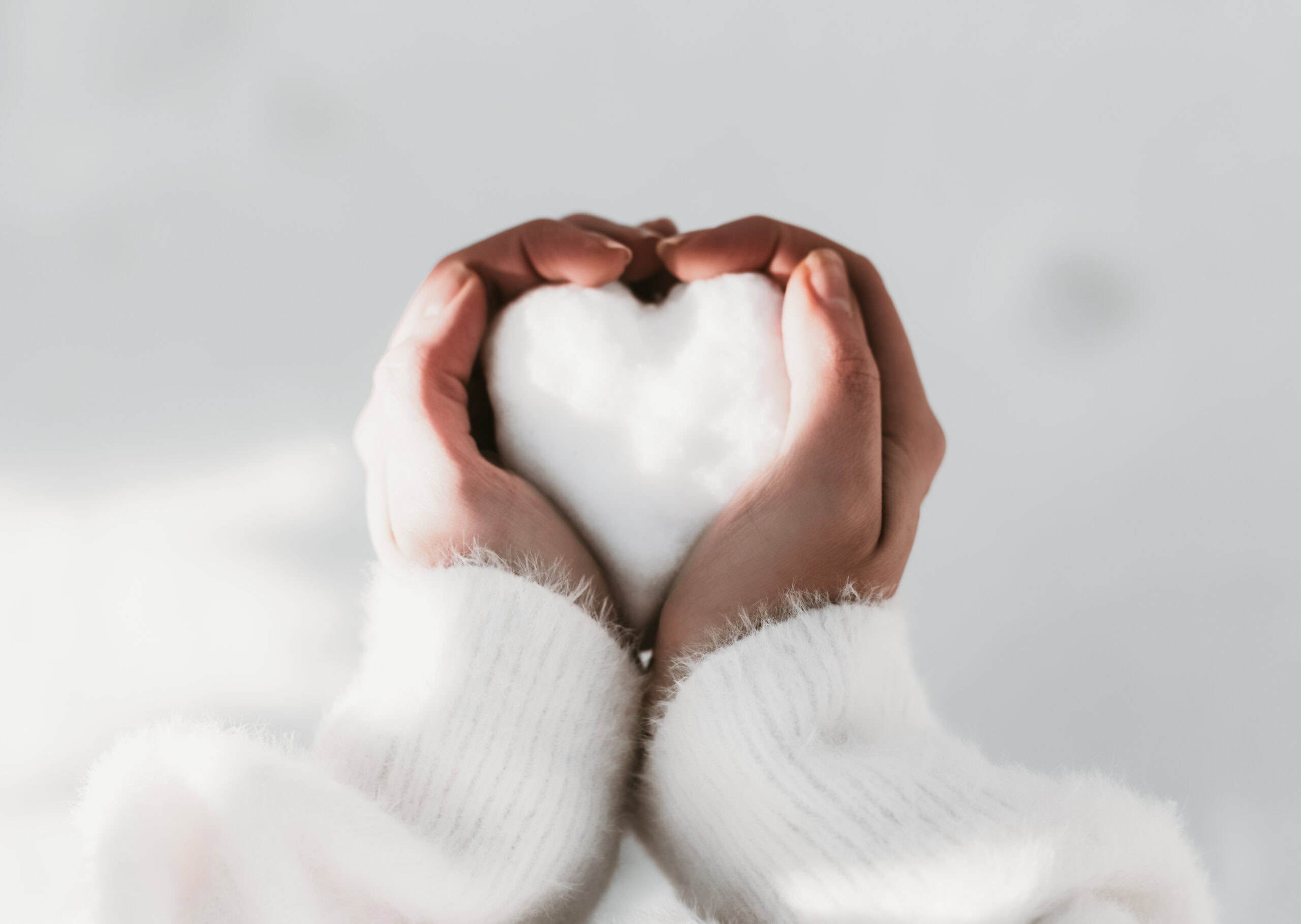 Hands in white sleeves cradle a heart-shaped snowball against a light background, capturing the joy of the holidays.