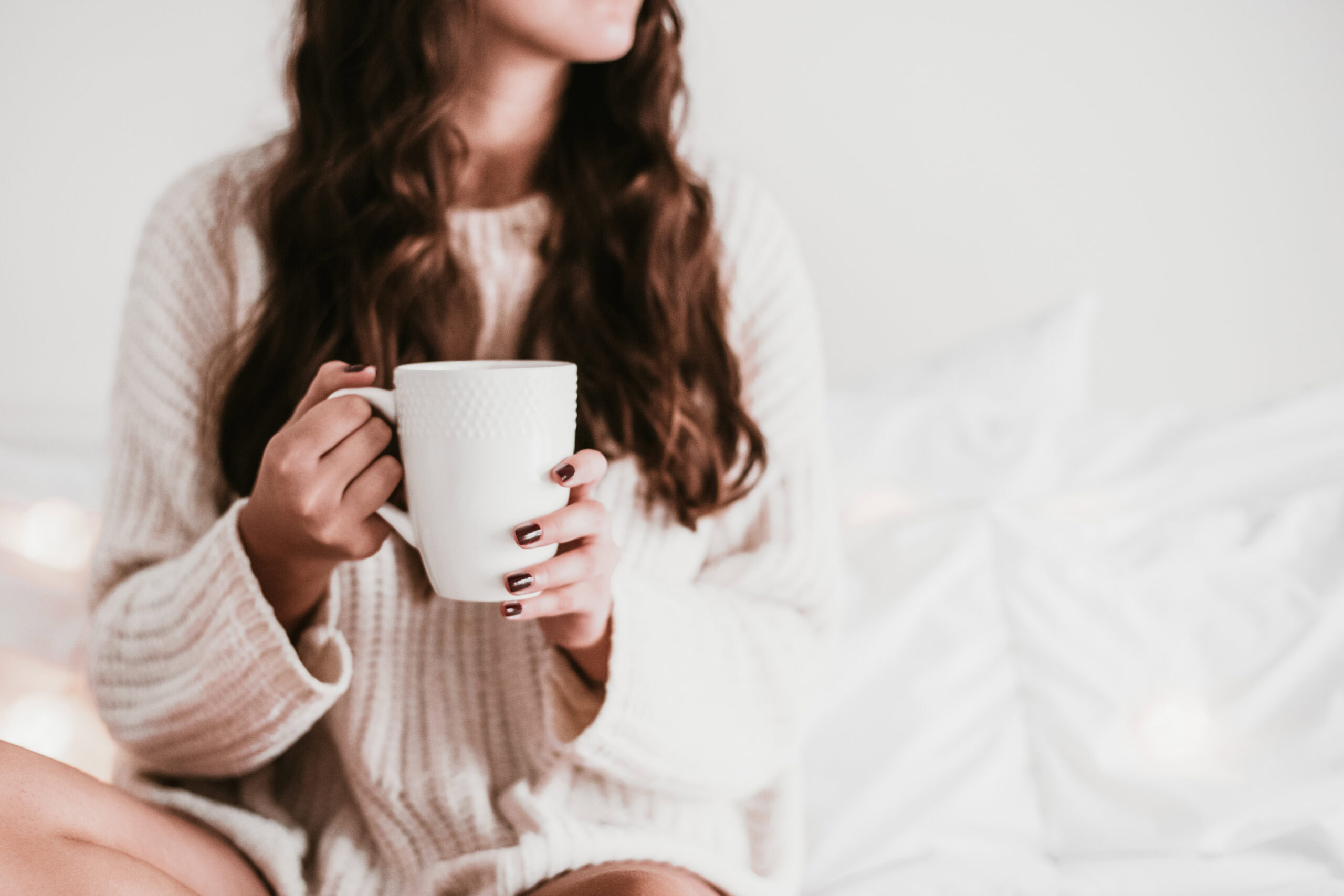 A person with long hair holds a large white mug, sitting on a bed wearing a cozy white sweater, finding solace in simple moments when loneliness persists.