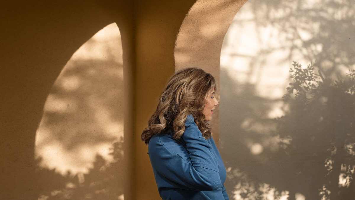 Carmen Hecox with wavy, healthier hair stands in front of a textured wall with arch-shaped shadows and dappled sunlight, wearing a blue top, facing sideways. Discover why heatless curls are the secret to fuller locks that radiate vitality and life.