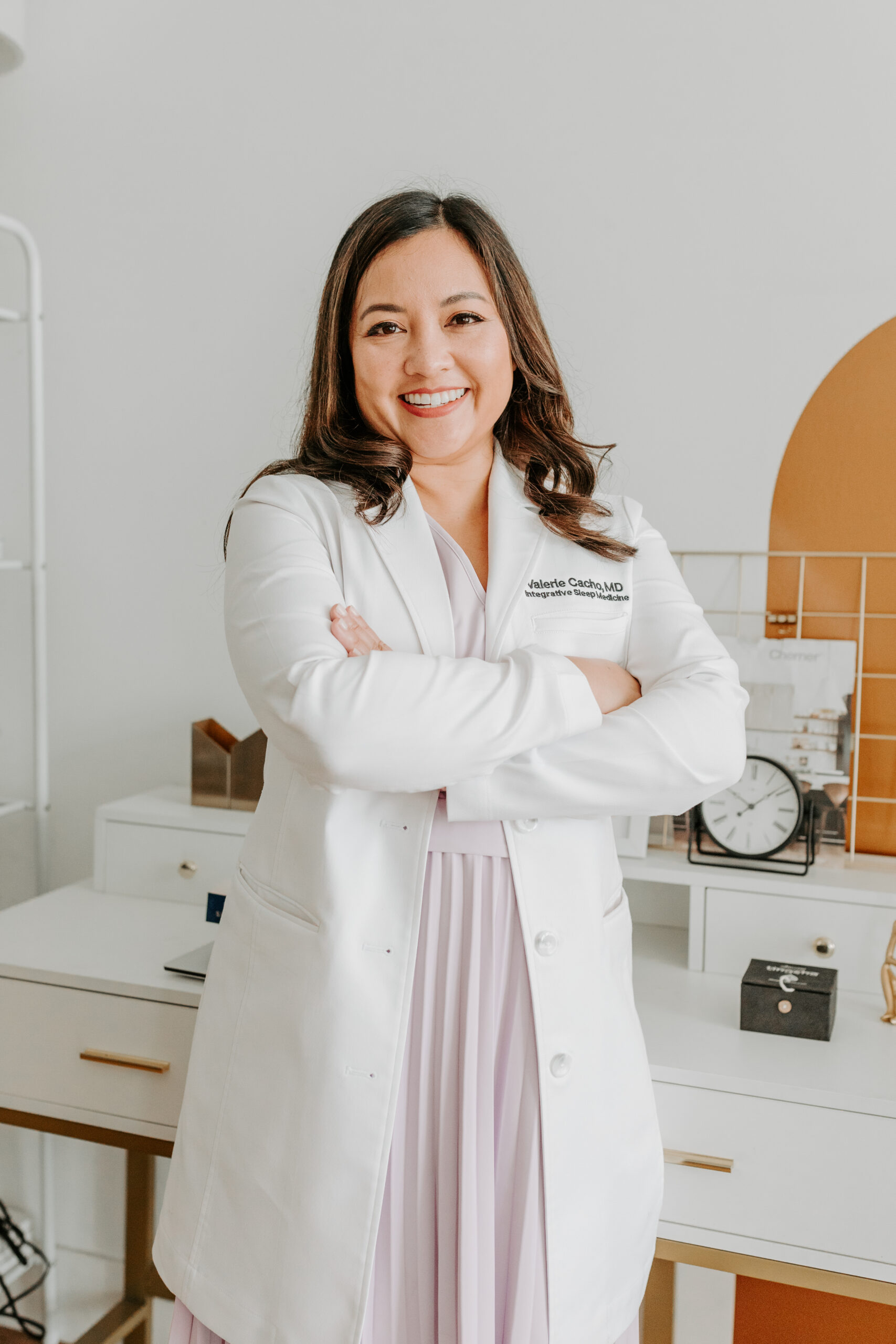 Dr. Val Cacho in a white coat stands confidently with arms crossed, embodying expertise in recognizing sleep apnea in women within an office setting.