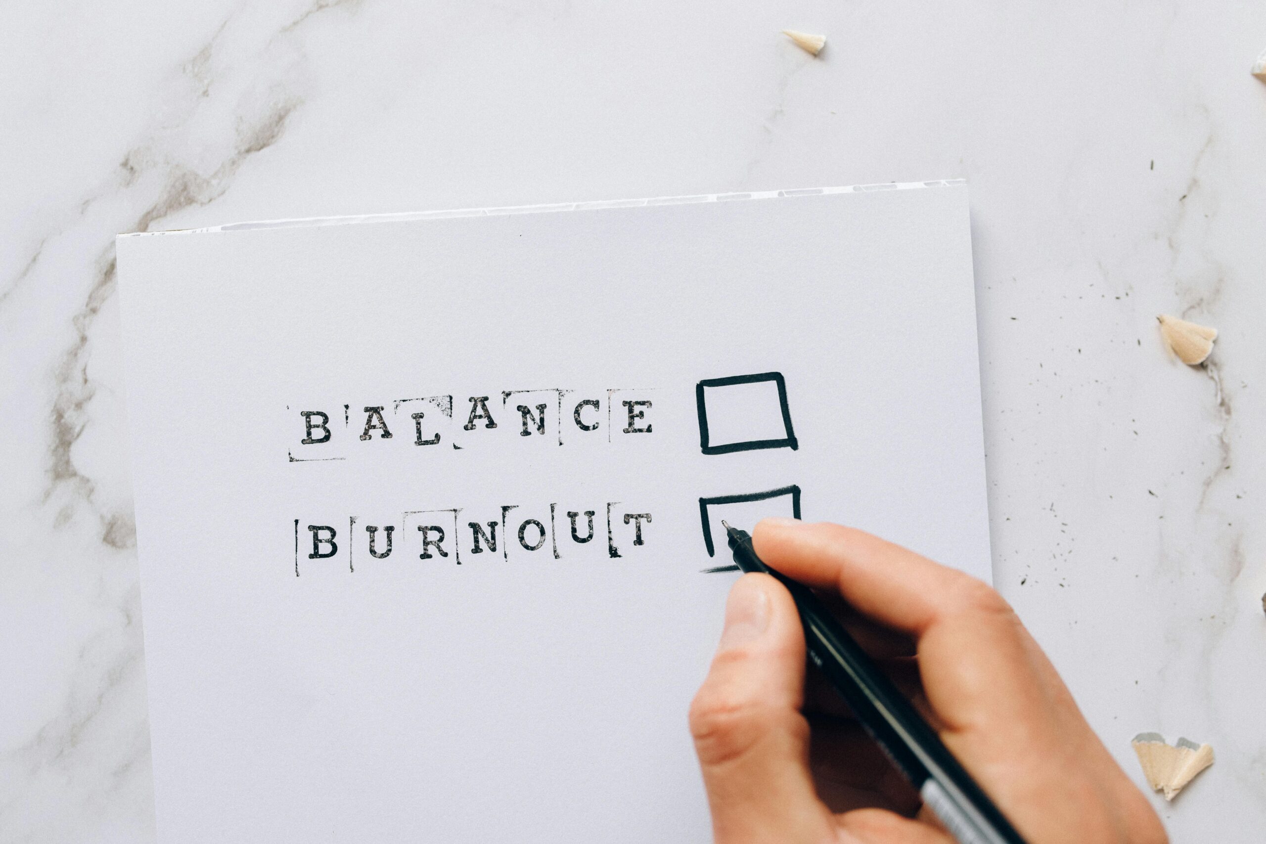 A hand grips a pen, marking the box beside “BURNOUT” on a paper, while “BALANCE” remains unchecked. Resting on a marble surface, this scene underscores the elusive art of balancing family duties, career ambitions, and self-care.