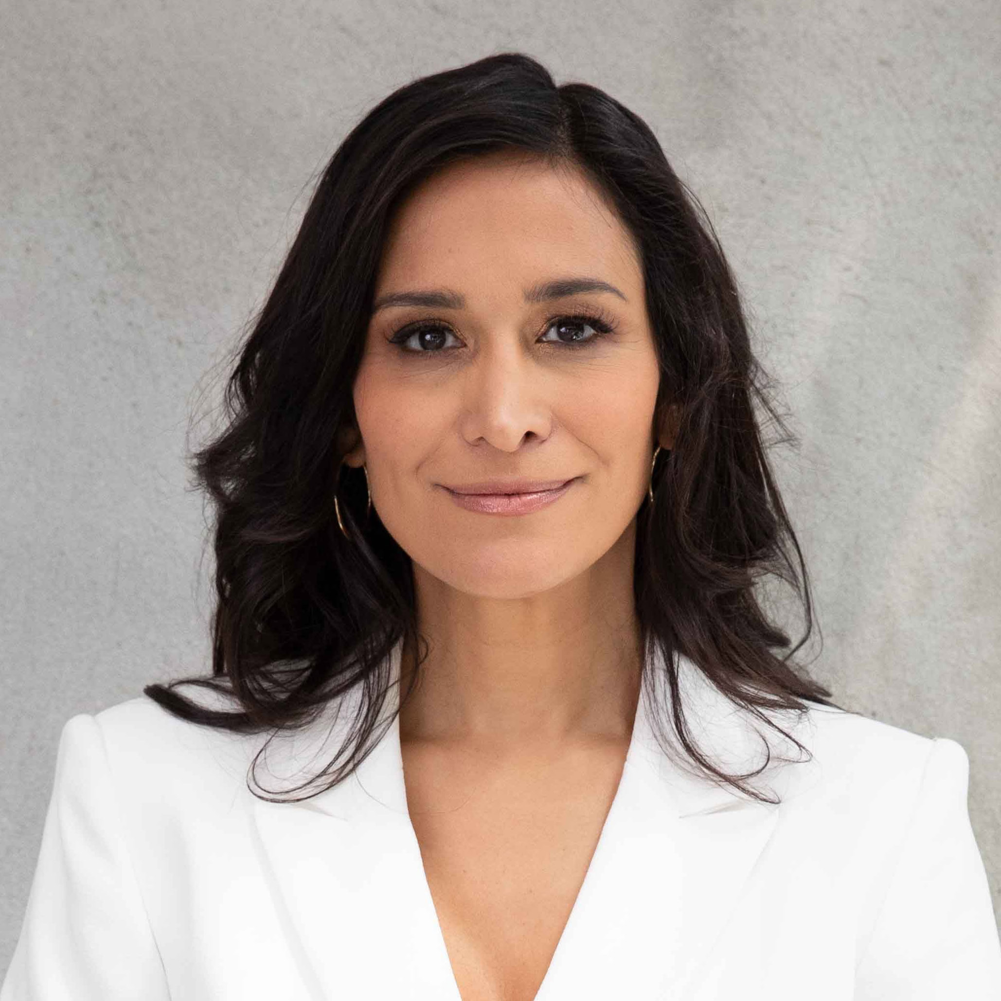 A woman with long dark hair and a white blazer smiles slightly against a gray background, embodying the grace of an upcoming guest.