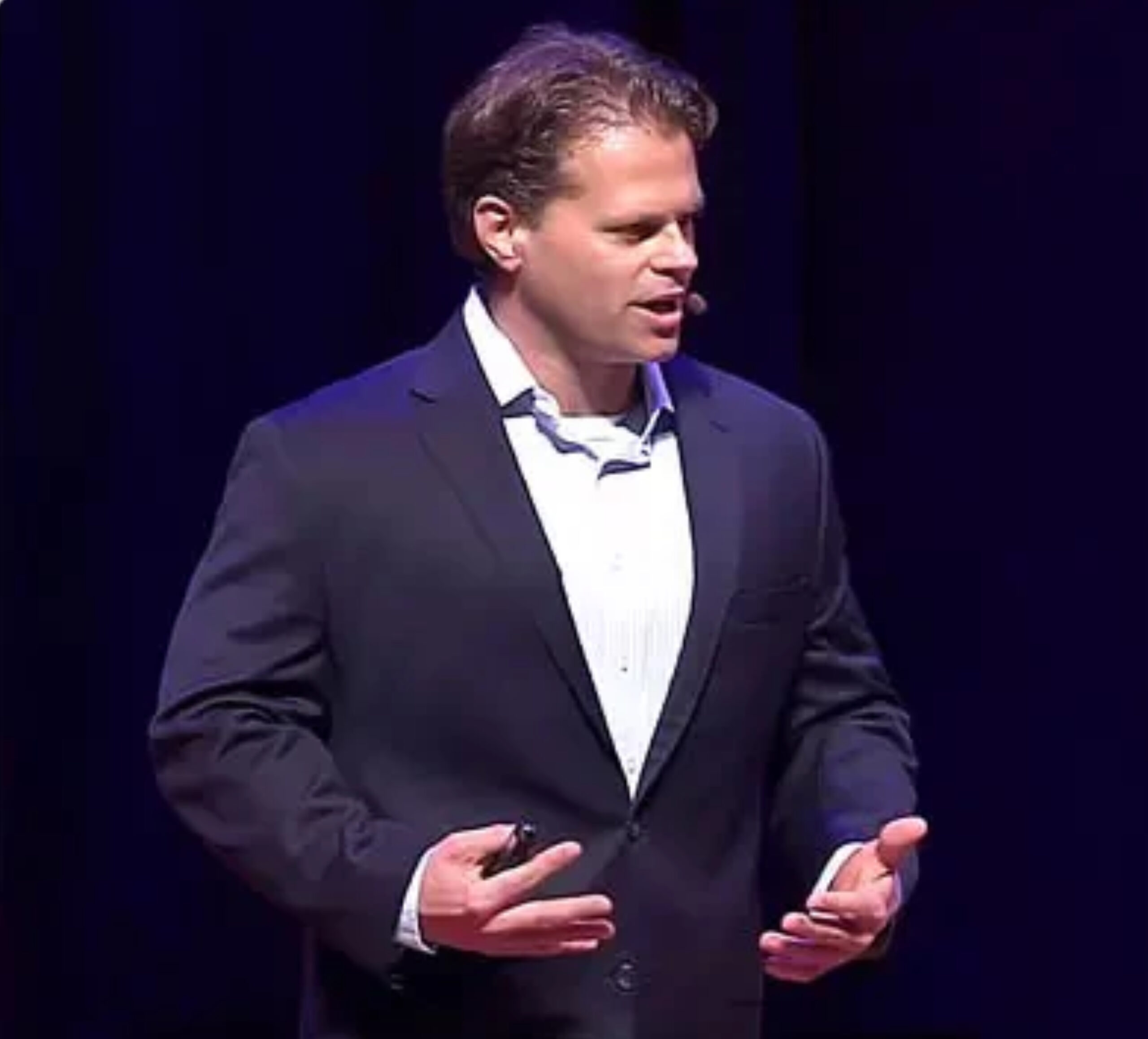 In a dimly lit setting, Dr. John Lewis in a suit speaks passionately on stage, sharing three tips to make your New Year's resolution work, all while wearing a headset microphone.