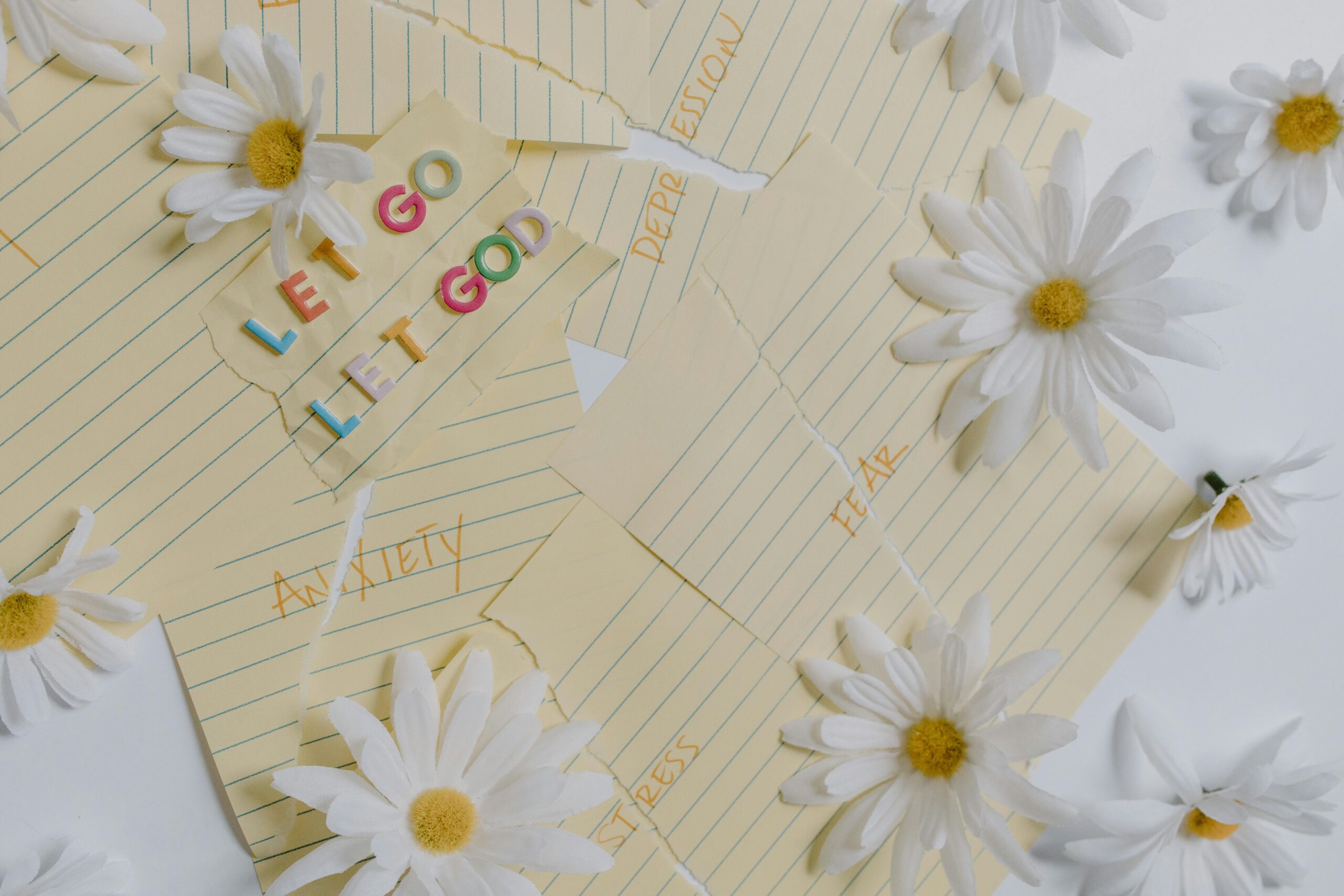 Torn lined paper labeled with words like "fear" and "stress" is surrounded by daisies, while colorful letters spell out "Let Go, Let God," embodying the theme of Embracing Faith for a Fulfilled 2025.