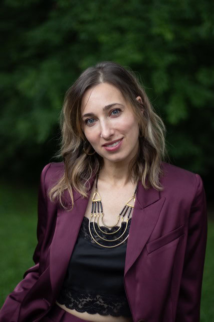 Upcoming Guest's Evalee Gertz with long hair in a purple suit and black lace top stands outdoors, exuding empowerment as she gracefully poses against a backdrop of lush green foliage.