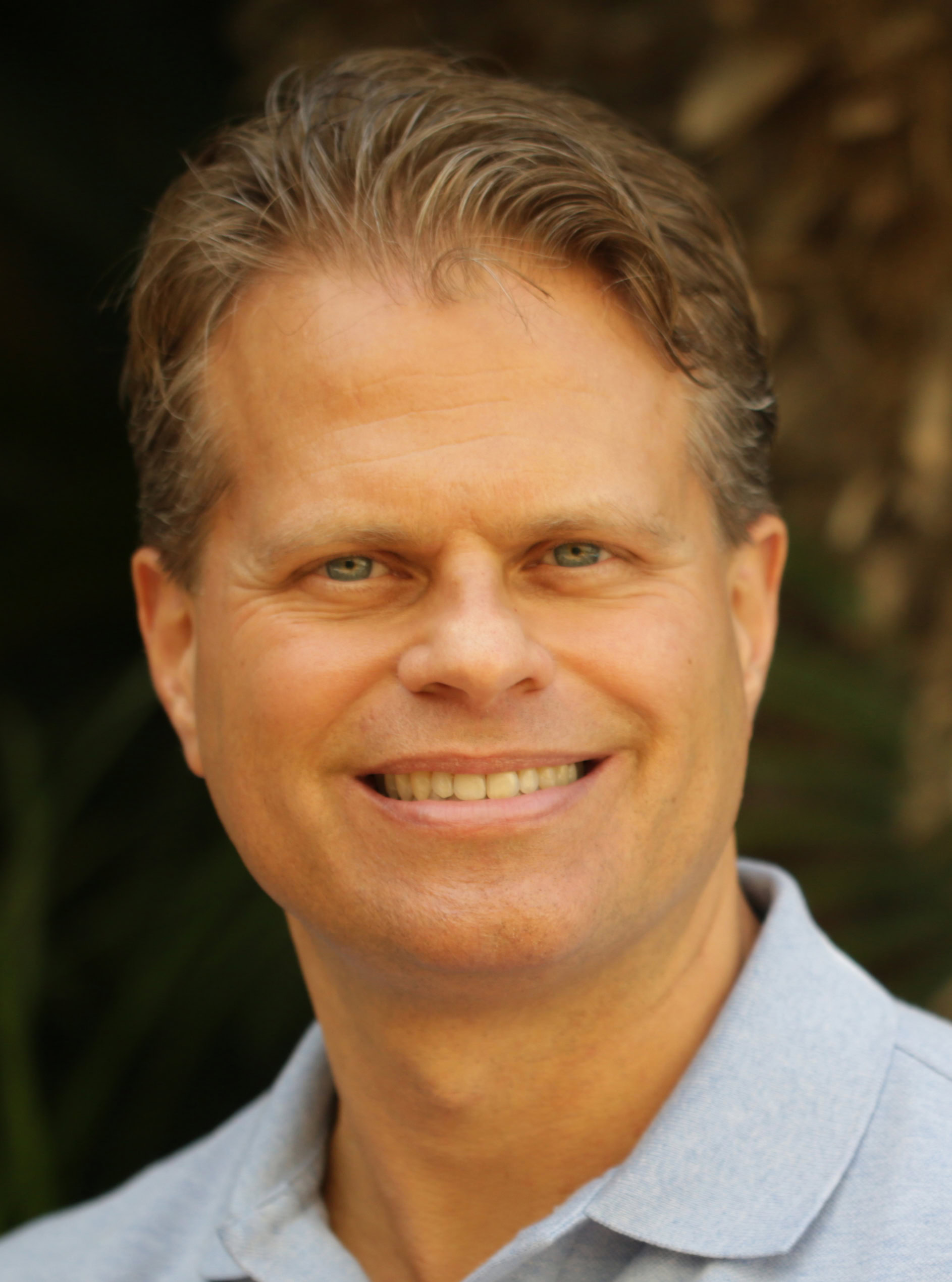 A smiling person with short, light brown hair embodies a sense of wellness in a light blue collared shirt against a blurred background.